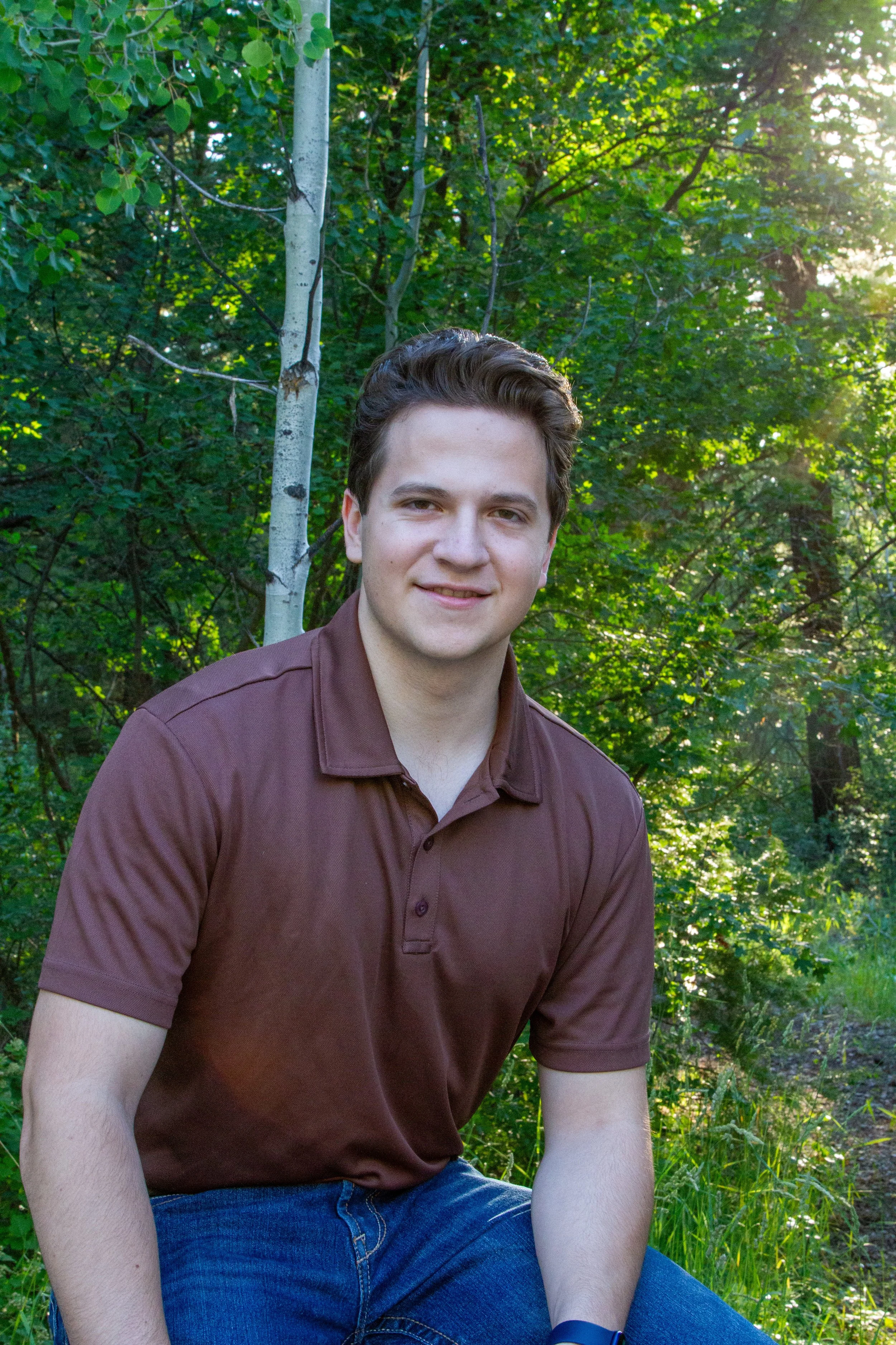 A young man with dark hair and fair skin smiling, sitting outdoors in a forest with green leaves and trees, wearing a brown polo shirt and blue jeans.