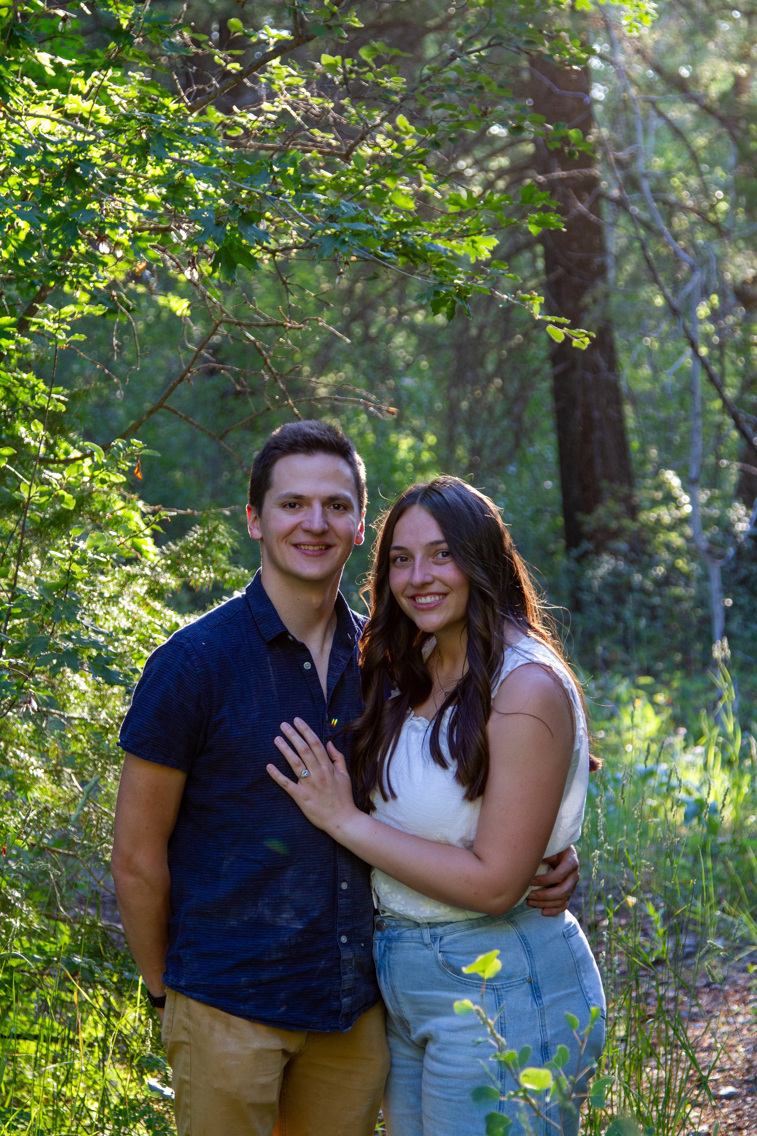 A young couple smiling and standing close together outdoors in a lush green forest during daylight, with sunlight filtering through the trees behind them.