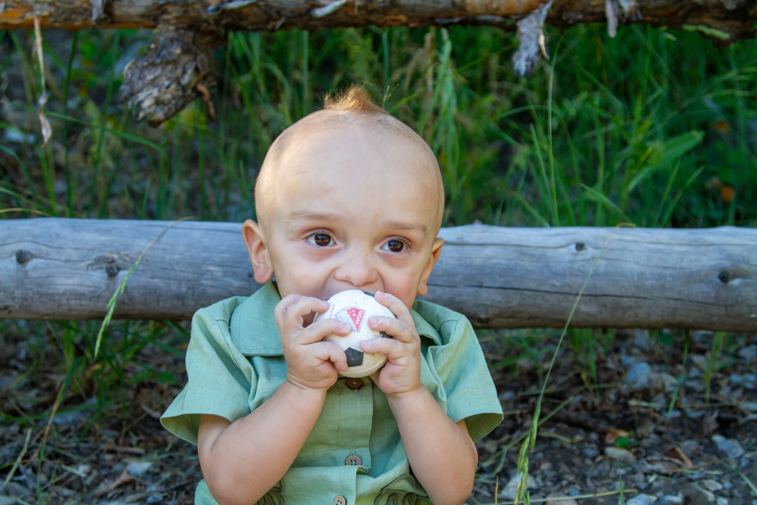 A young boy with a bald head and green shirt sitting on a wooden bench outdoors, holding a soccer ball and drinking from a bottle, with green grass and plants in the background.