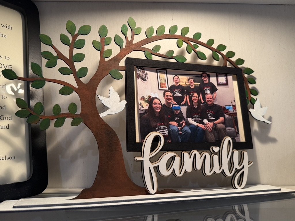 Decorative wooden family tree with green leaves and white dove cutouts, next to a framed family photo showing a group of people, with a white 'family' sign at the bottom.