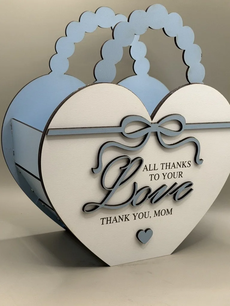 A decorative wooden box shaped like a heart, painted in white and light blue, with small cloud-like handles on top. The front has a ribbon and bow design with the text 'All thanks to your love. Thank you, Mom' and a small heart icon at the bottom.