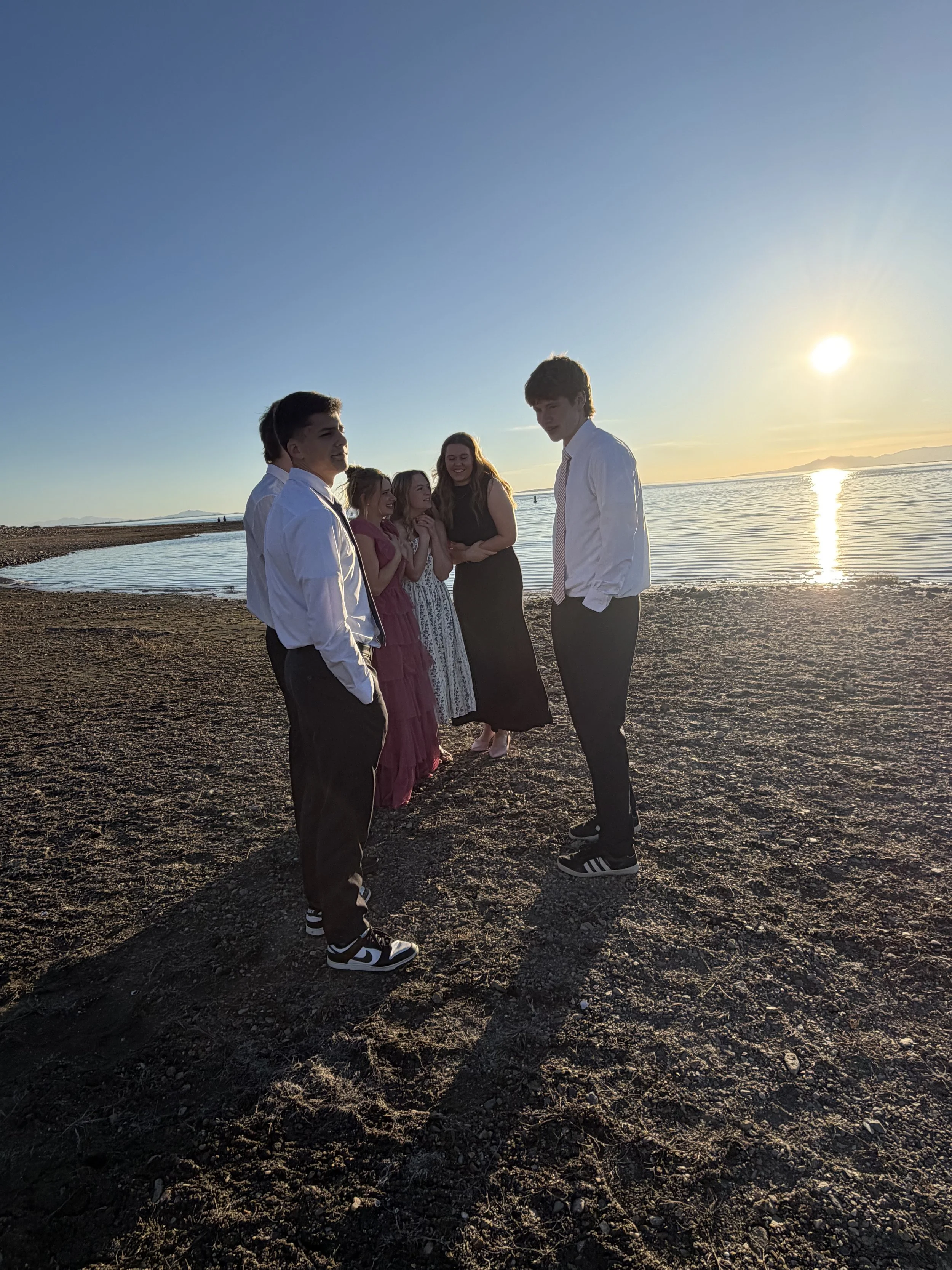 Group of six people standing on a pebble beach during sunset, engaged in conversation. The water is calm, reflecting the setting sun, with distant mountains on the horizon.