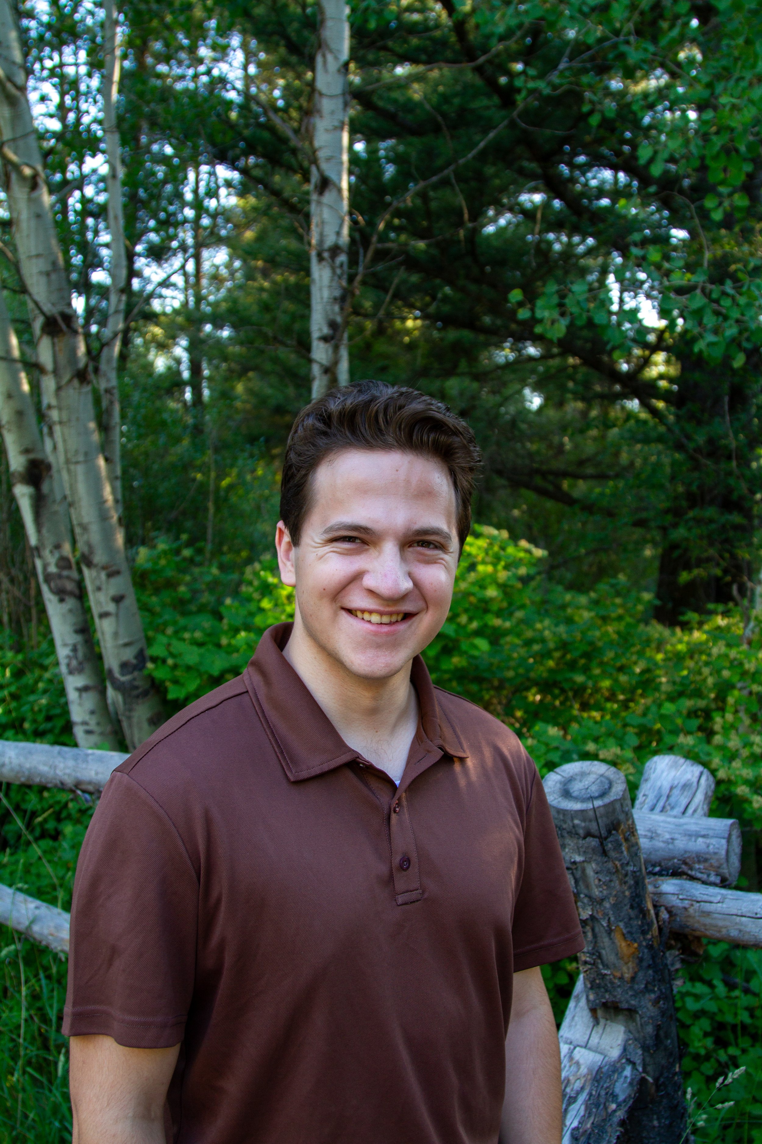 A young man with dark hair smiling outdoors, wearing a brown polo shirt, in a green wooded area with trees and a rustic wooden fence.