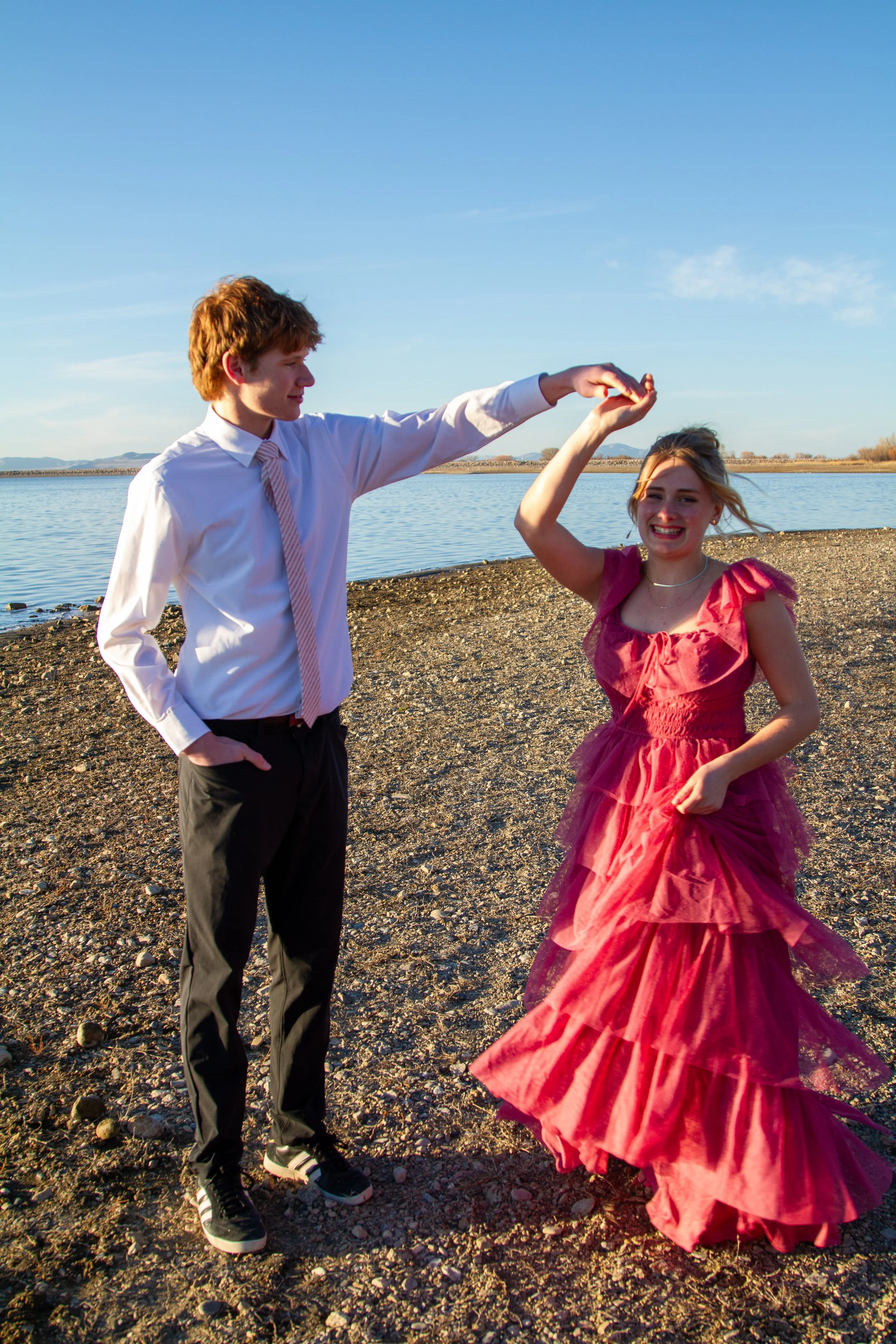 A young man in formal attire holding the hand of a woman in a pink gown dancing by a body of water during sunset.