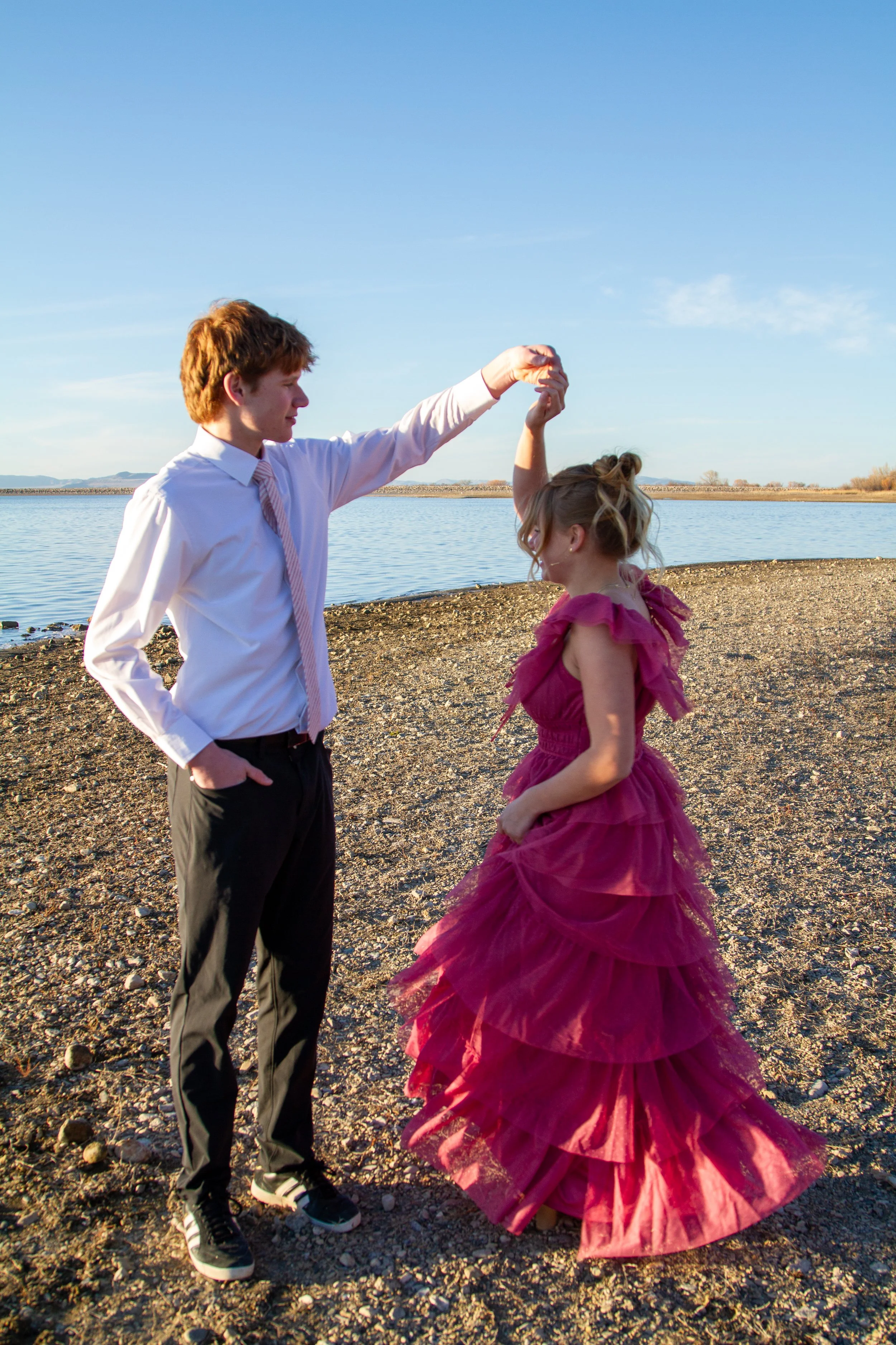 A young man and woman dancing on a rocky beach by a lake, with the man holding the woman's hand as she twirls in a pink ruffled dress, under a clear blue sky.