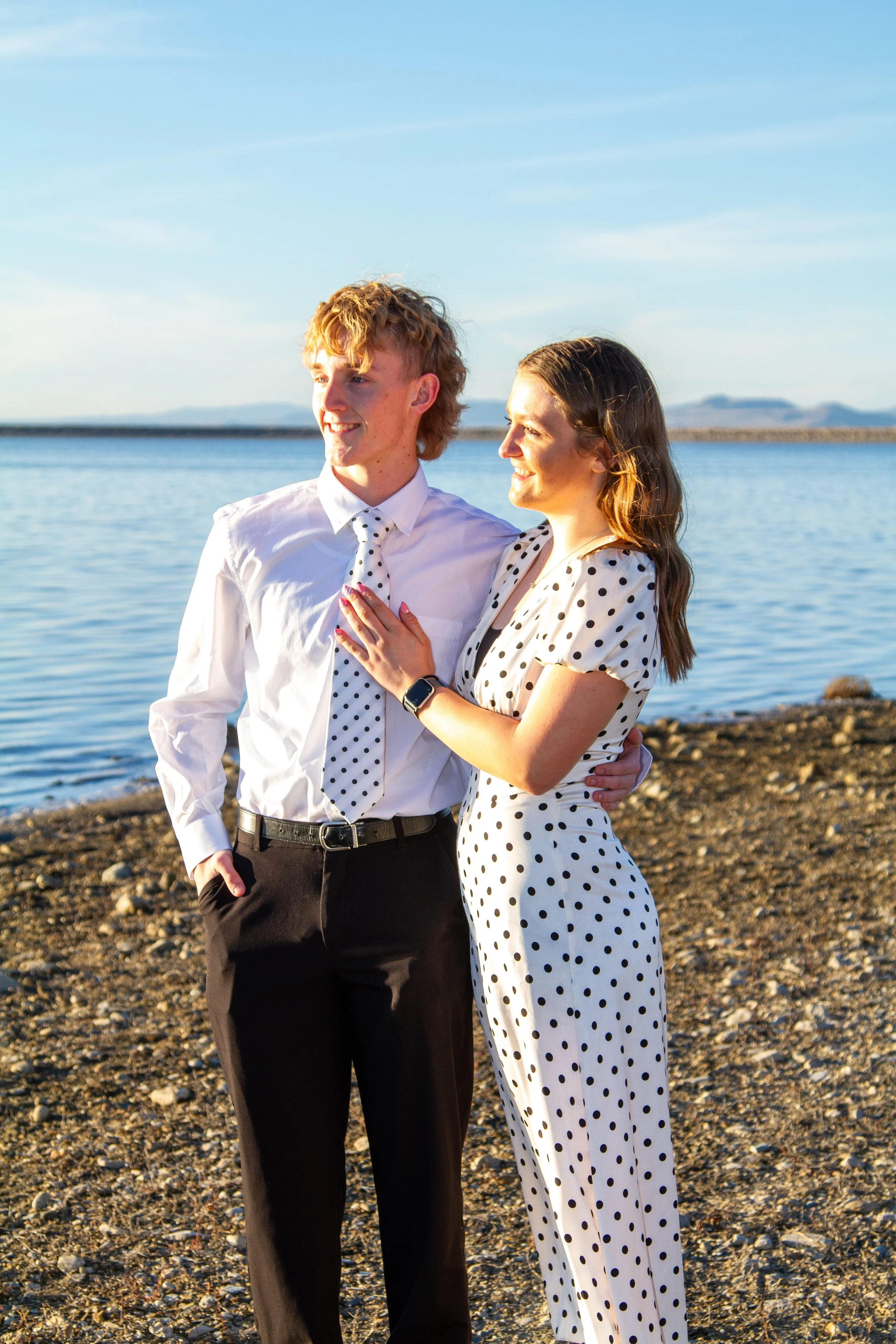 A young couple standing on a rocky beach by the water, smiling and looking into the distance, dressed in matching polka dot outfits with a sunny sky in the background.