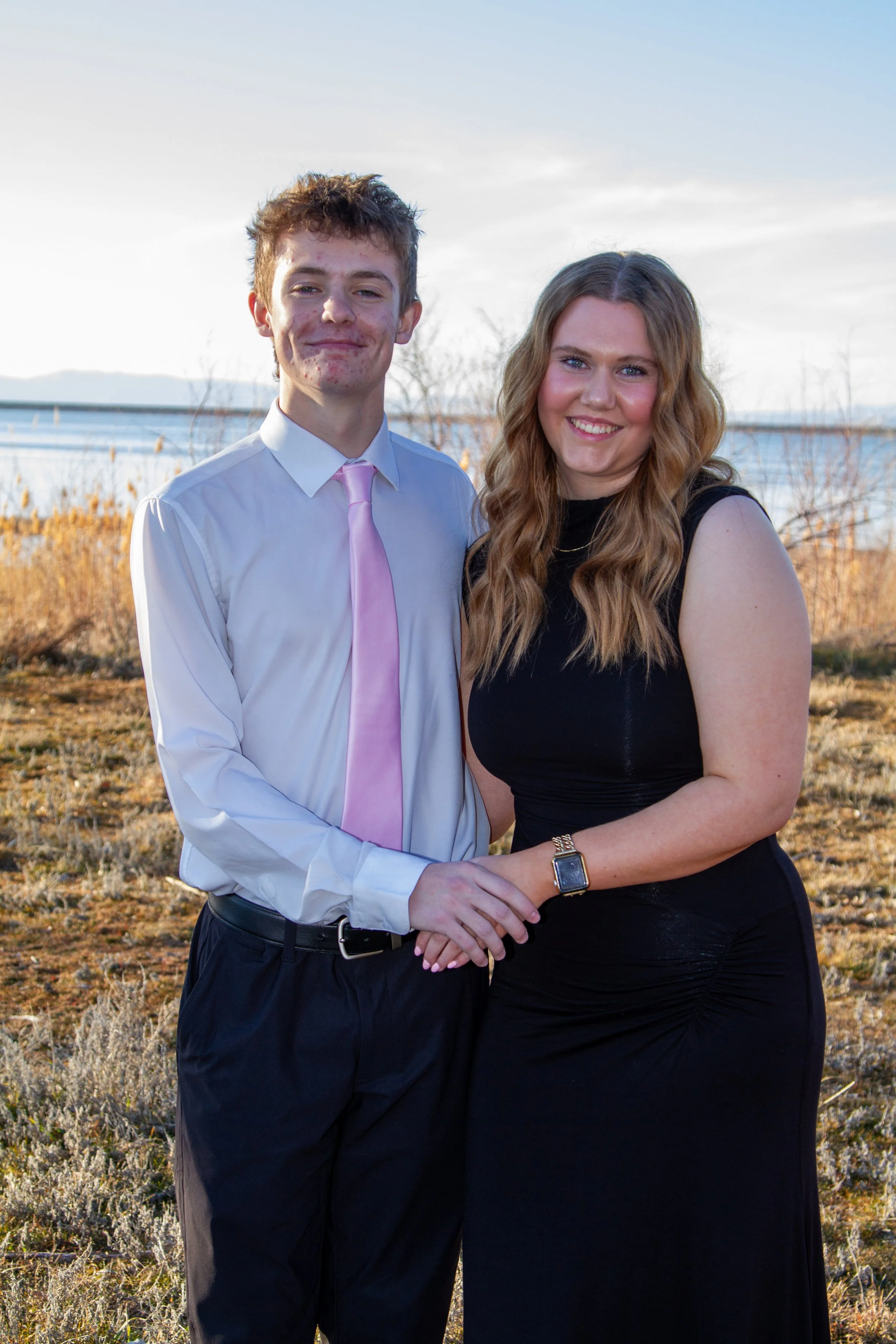 A young man and woman standing outdoors near a body of water, holding hands and smiling at the camera, with autumn foliage and clear skies in the background.