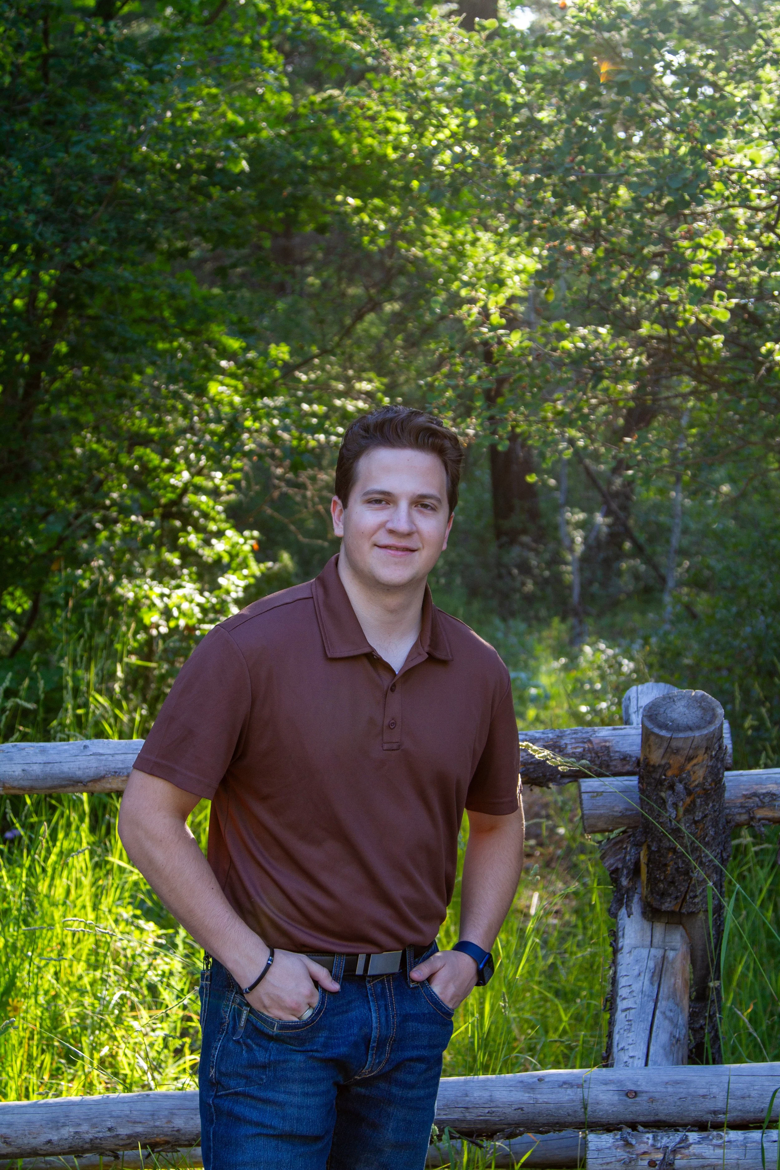 A young man with dark hair wearing a brown polo shirt and blue jeans standing outdoors next to a wooden fence with greenery and trees in the background.