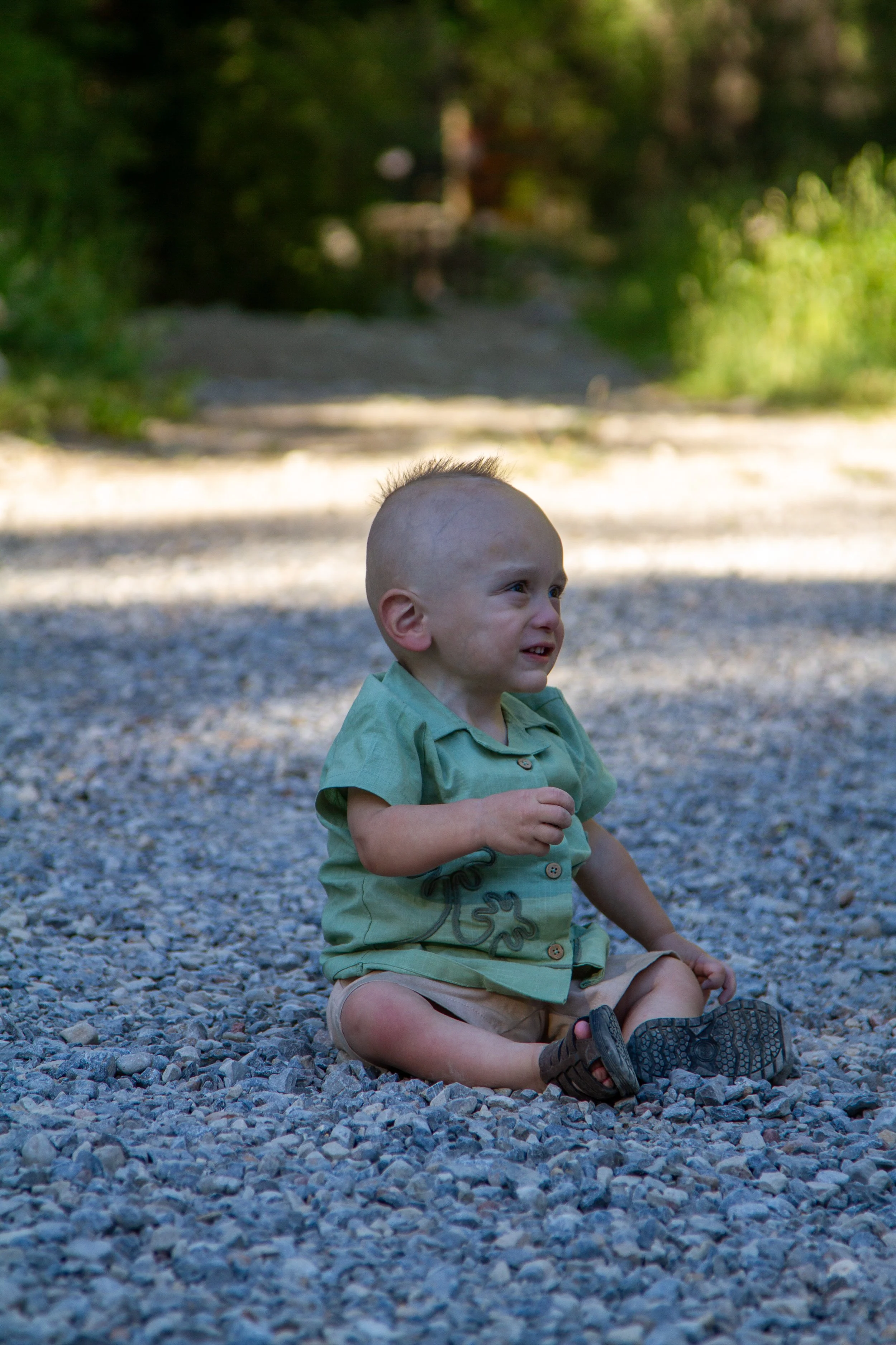 A young child with a mohawk hairstyle, sitting on a gravel path outdoors, smiling and looking to the side, surrounded by green trees and bushes.