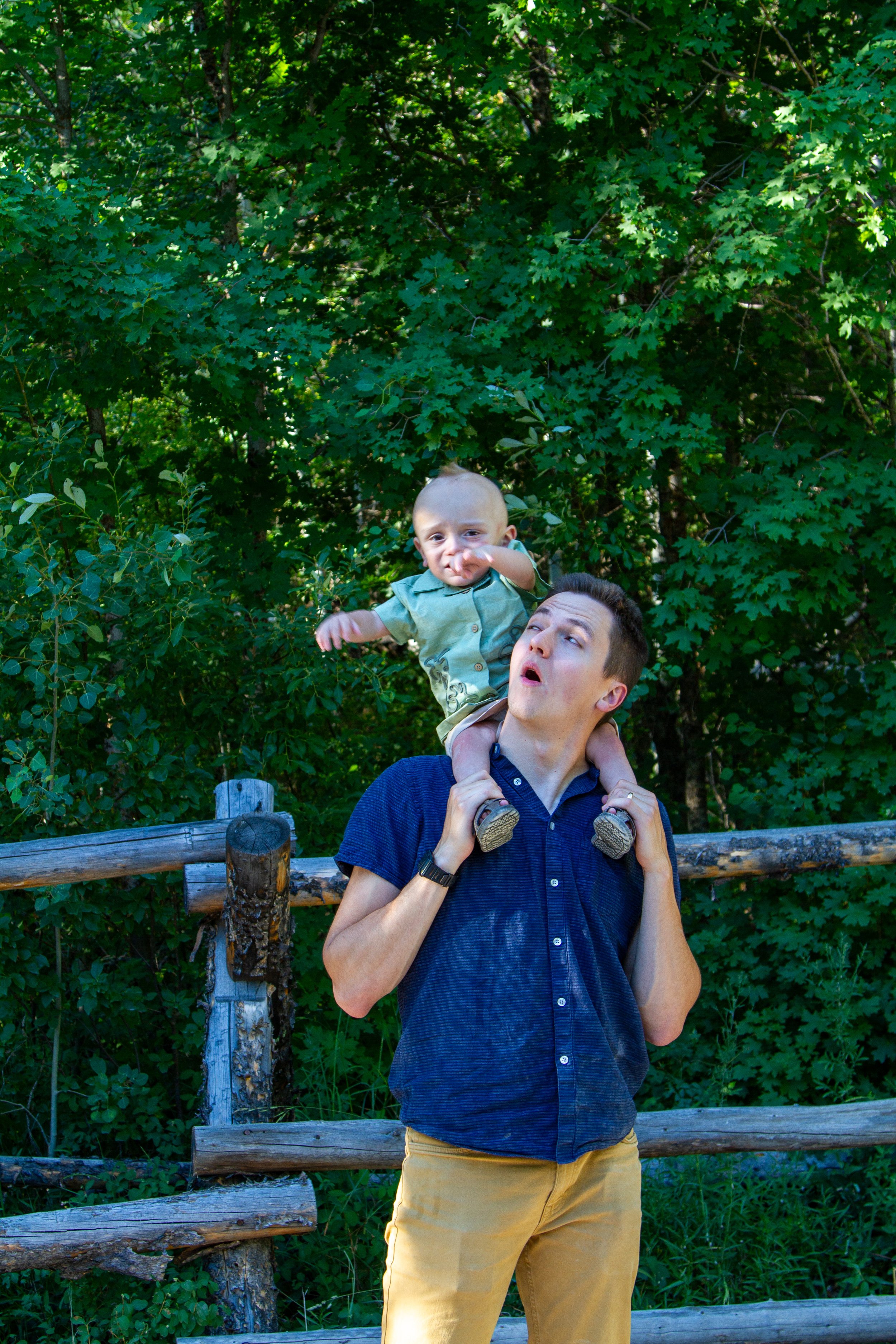 A man with short dark hair is holding a young boy with blonde hair on his shoulders in front of a green leafy background. The boy is reaching toward the camera with a curious expression, while the man looks slightly to the side with an open mouth.