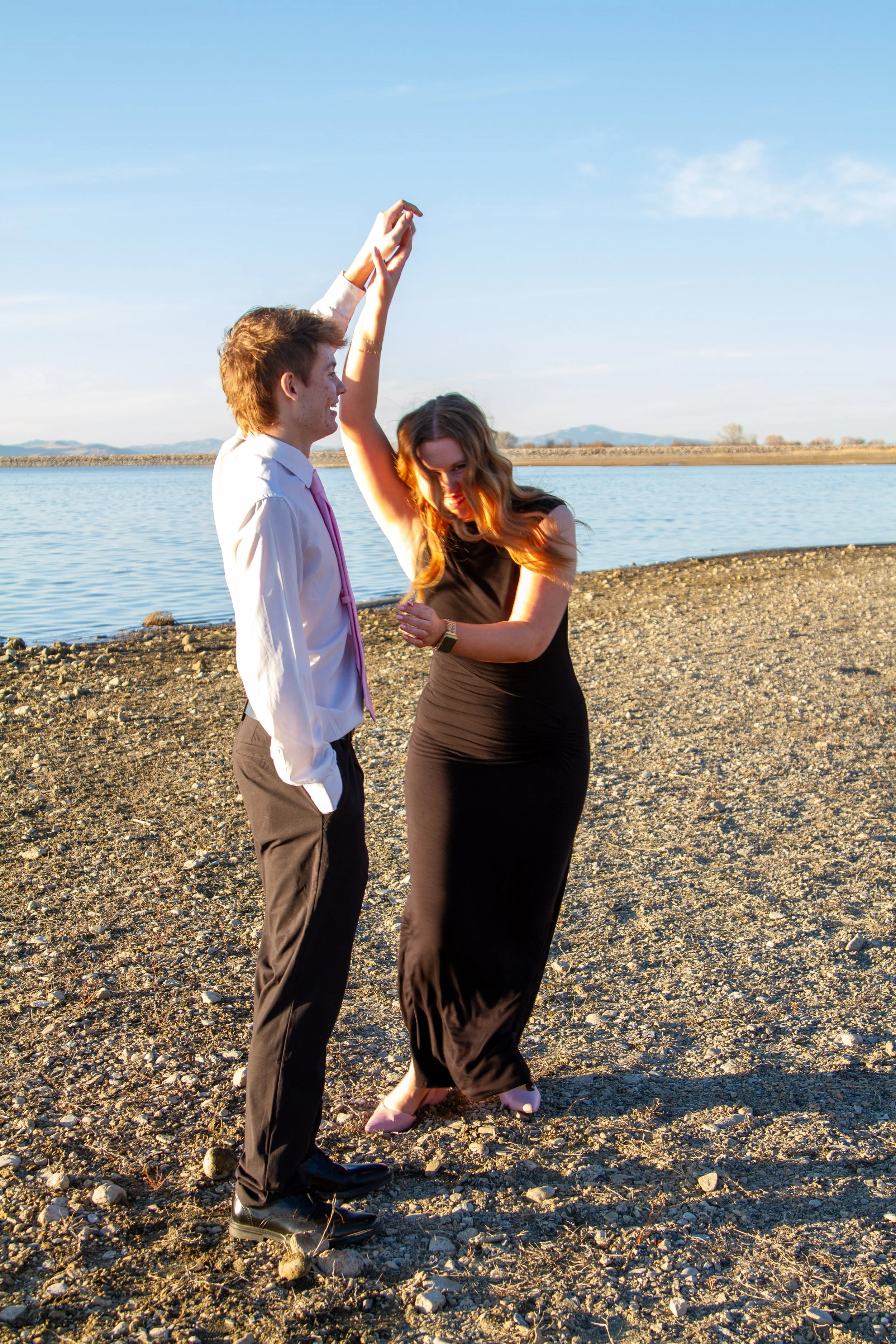 A man in dress pants, shirt, and tie dancing with a woman in a long black dress on a rocky shoreline near a body of water, with clear skies and distant mountains in the background.