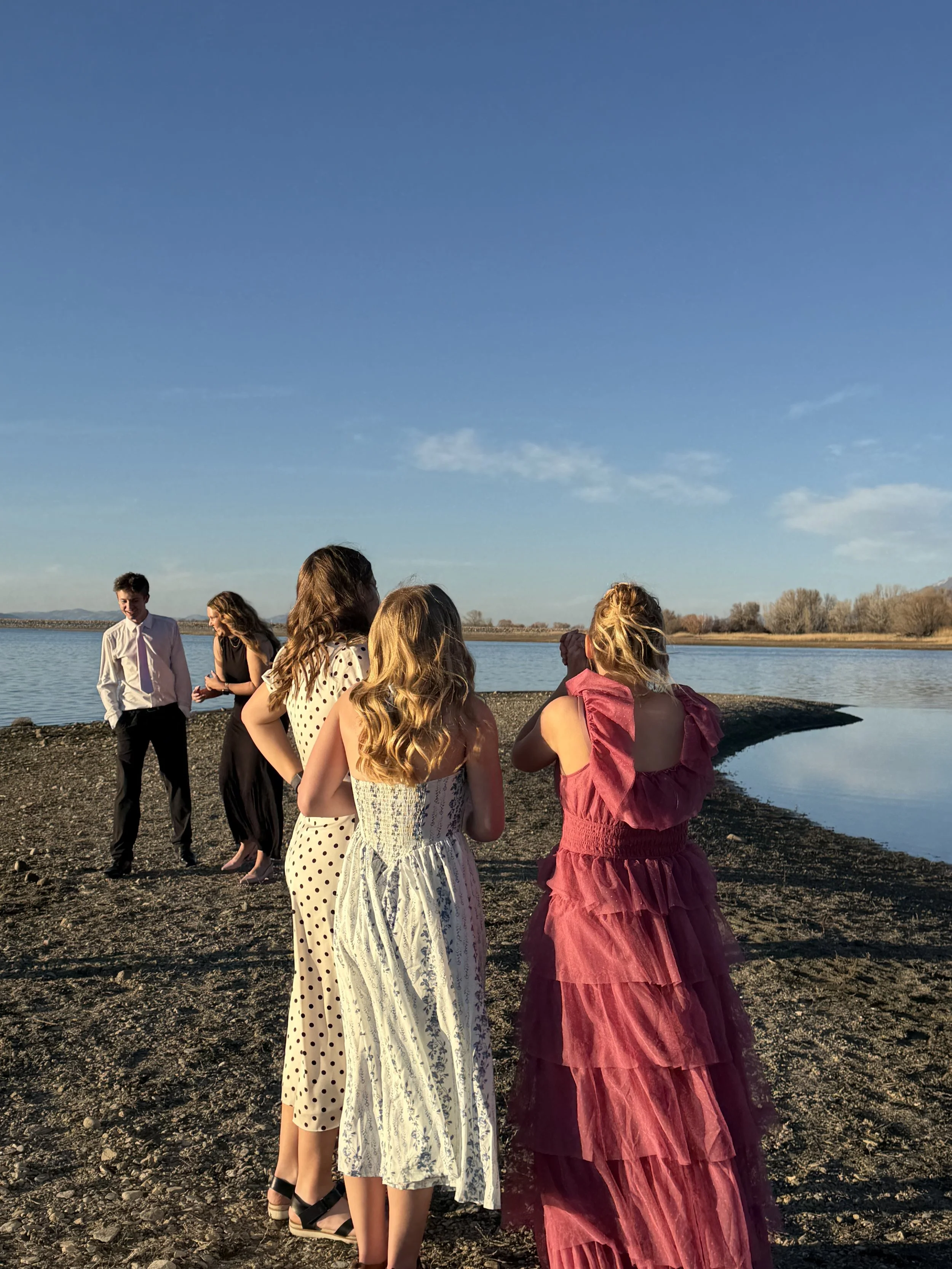 A group of six people, four women and two men, standing on a rocky shoreline by the water during sunset, dressed in elegant clothing.