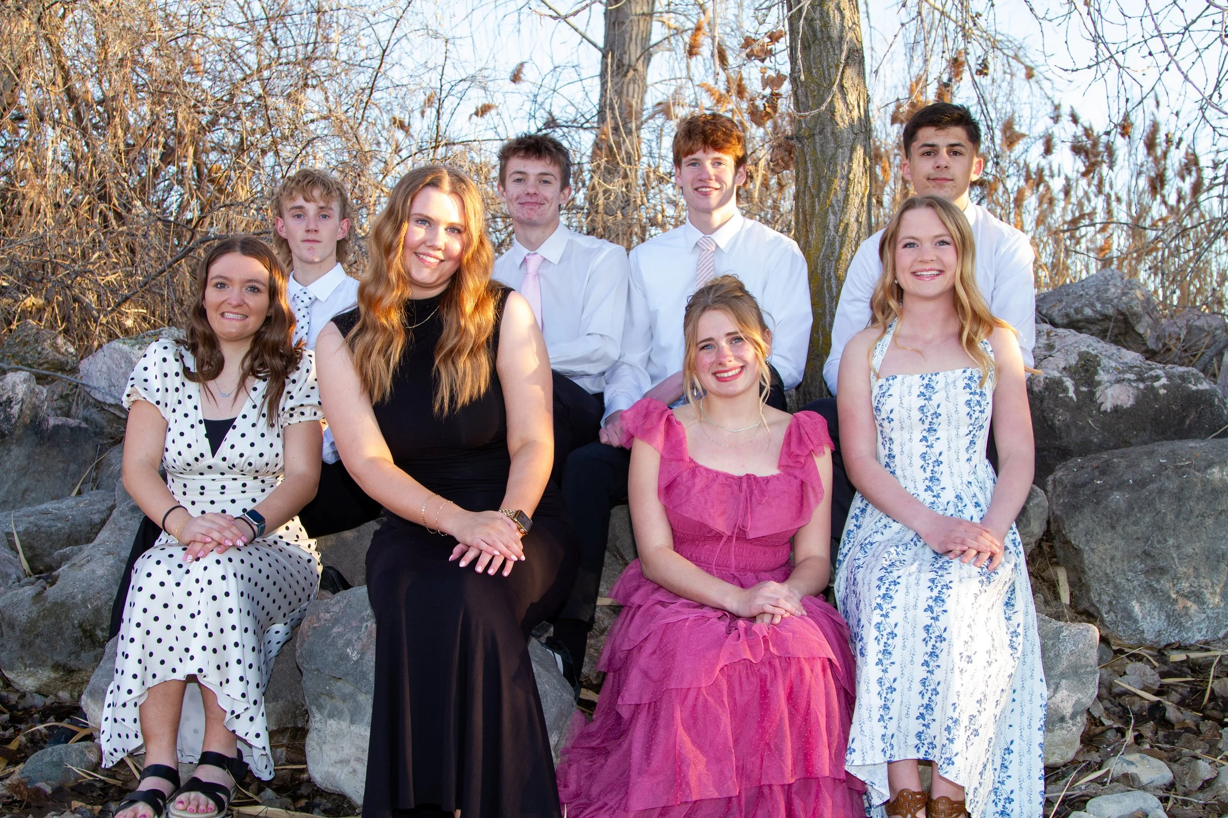 Group of ten young people, five women and five men, sitting and standing outdoors on rocks and branches, dressed in semi-formal attire, smiling at the camera, with trees and branches in the background.