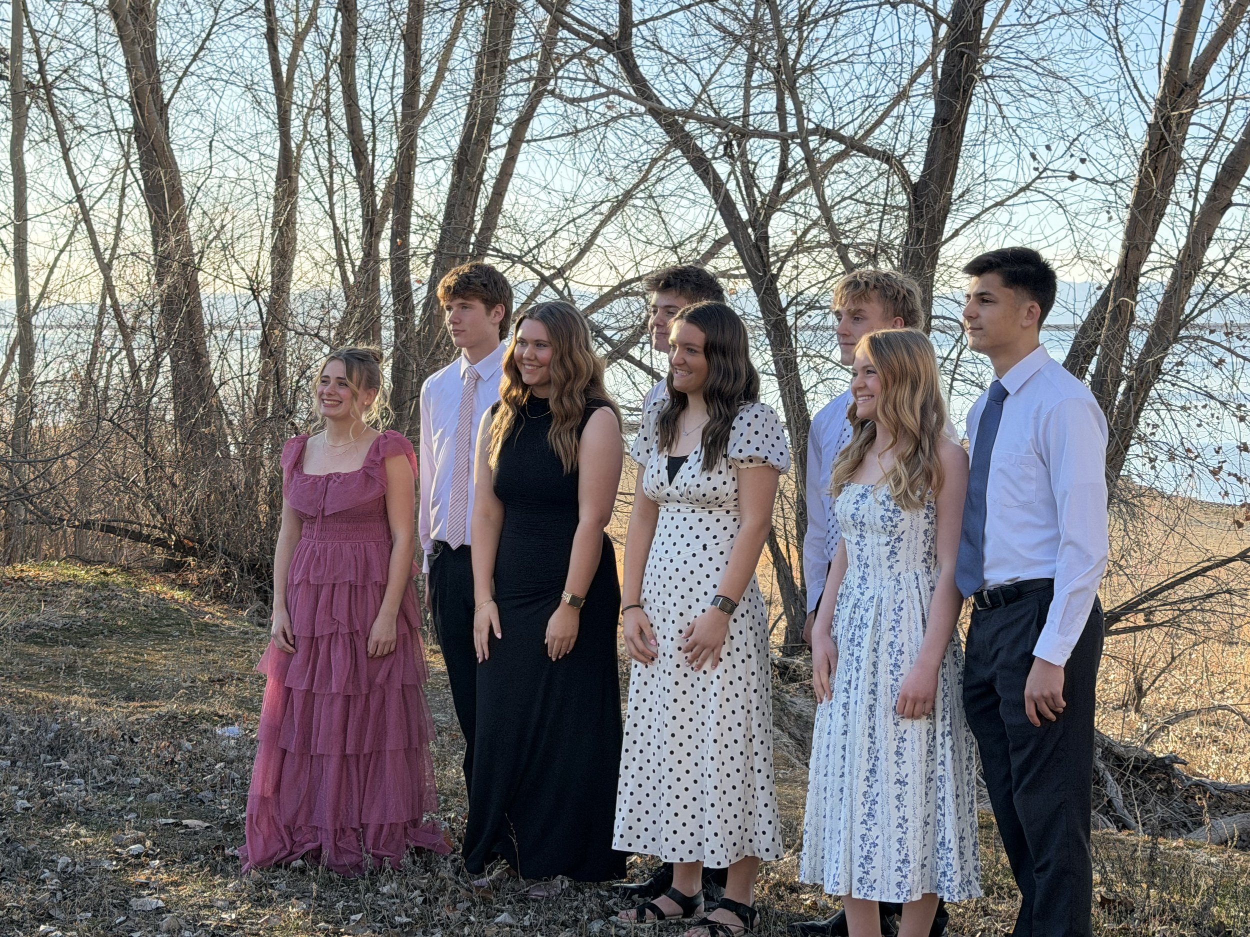 Eight teenagers standing outdoors near leafless trees, dressed in semi-formal attire, smiling and posing for a photo during sunset.
