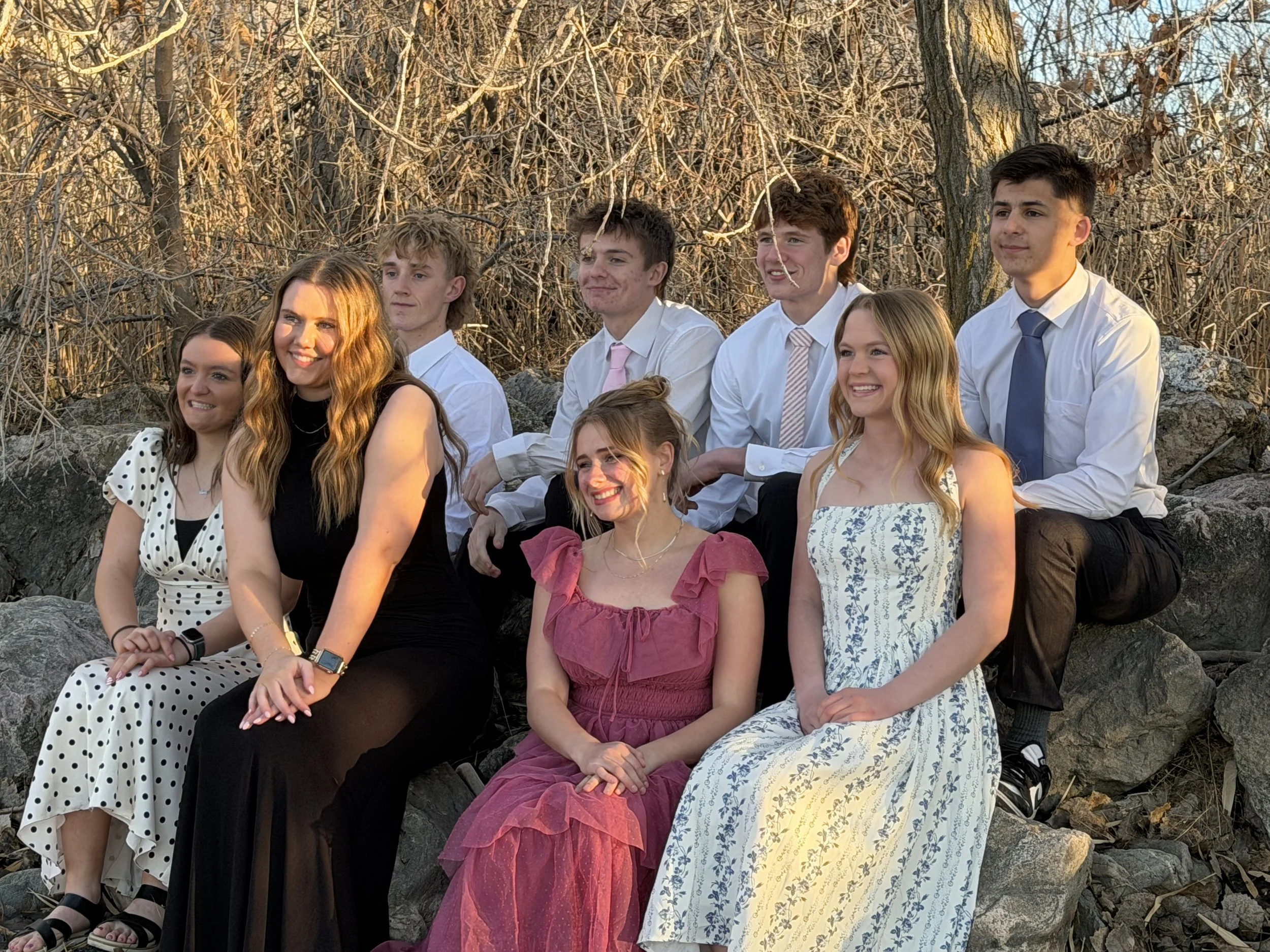 Group of nine young people outdoors on rocks with leafless trees in background, dressed semi-formally, smiling.
