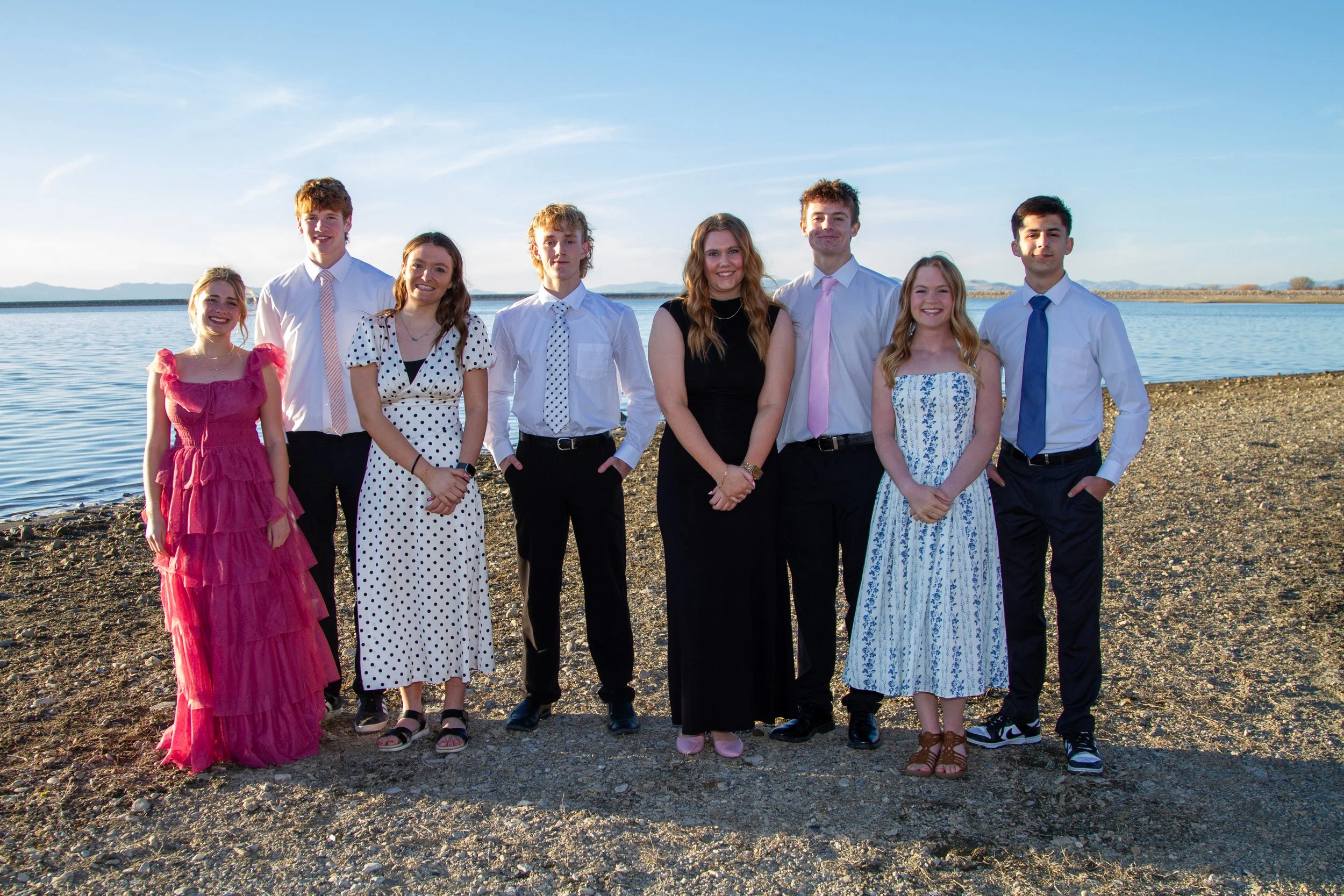 Group of nine young people standing on a rocky beach near a body of water, smiling and dressed in semi-formal attire with dresses and shirts with ties, under a clear blue sky.