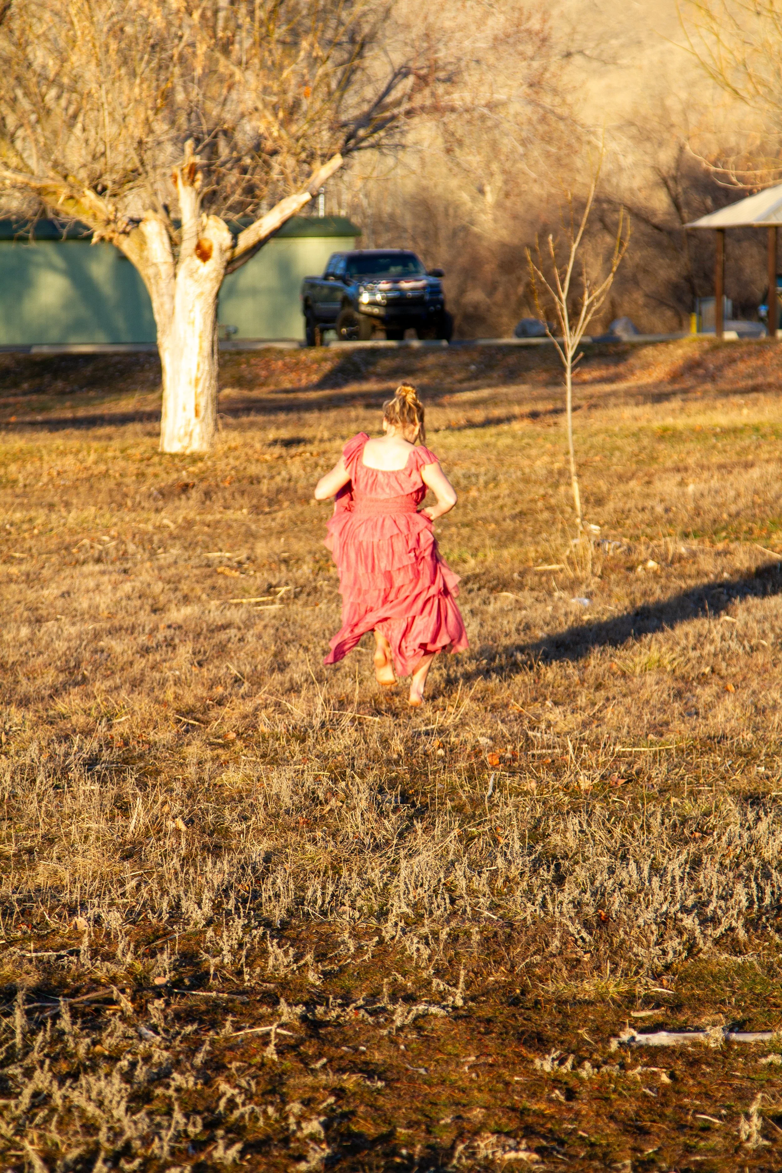 A girl in a pink dress running barefoot on a grassy field with trees and a parked vehicle in the background.