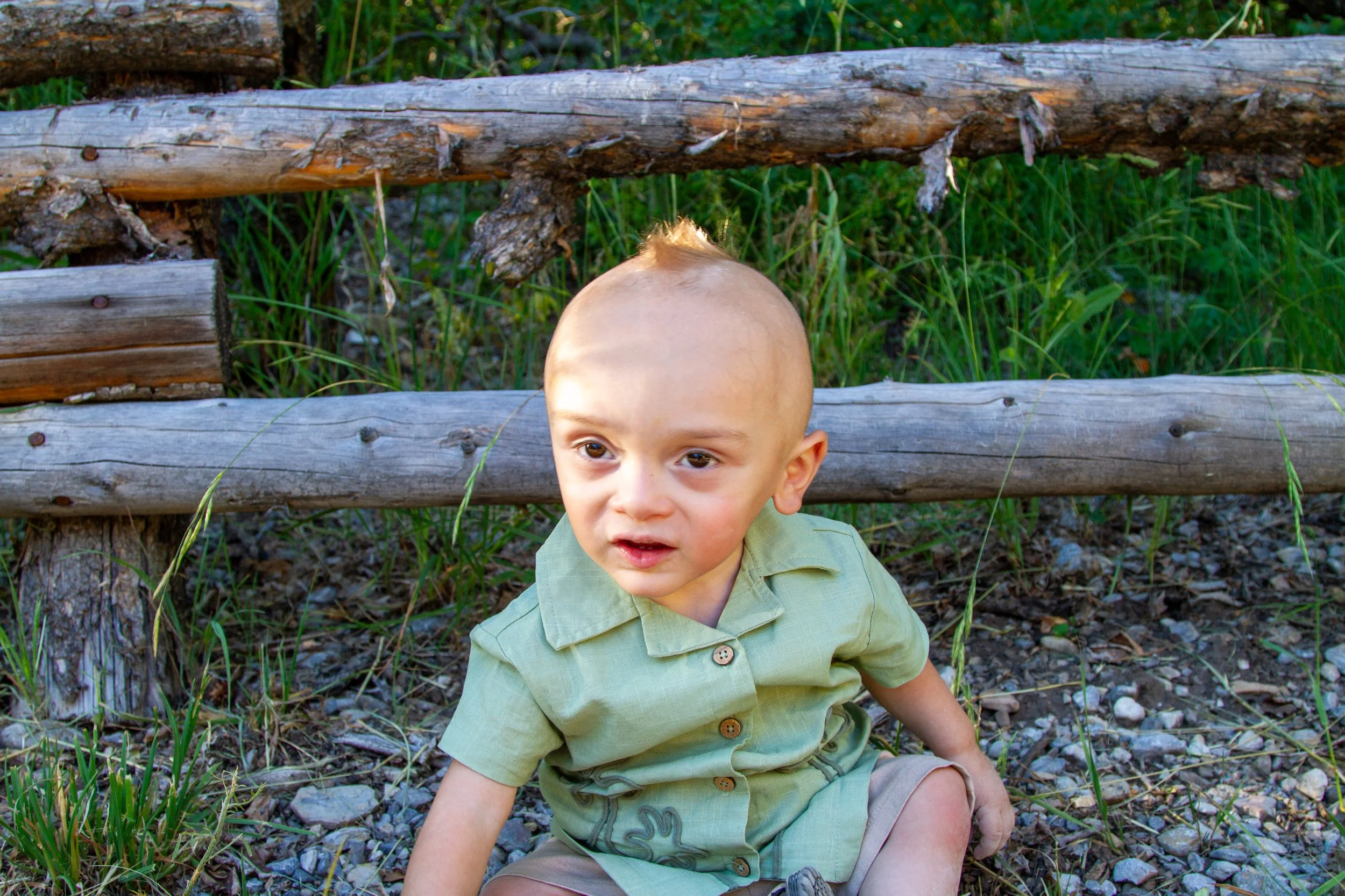 Young boy with a shaved head and wearing a light green shirt sitting in front of a weathered wooden fence outdoors with grass and gravel around him.