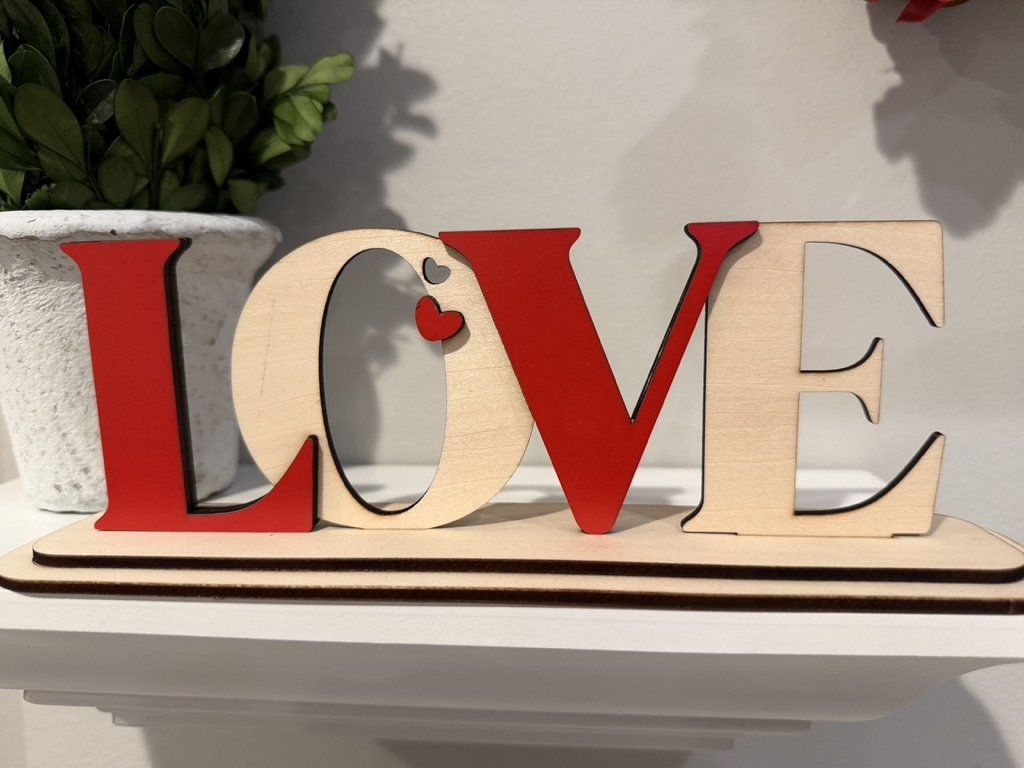 Decorative wooden and red metal letters spelling the word "LOVE" on a white shelf, with a plant in a white pot in the background.