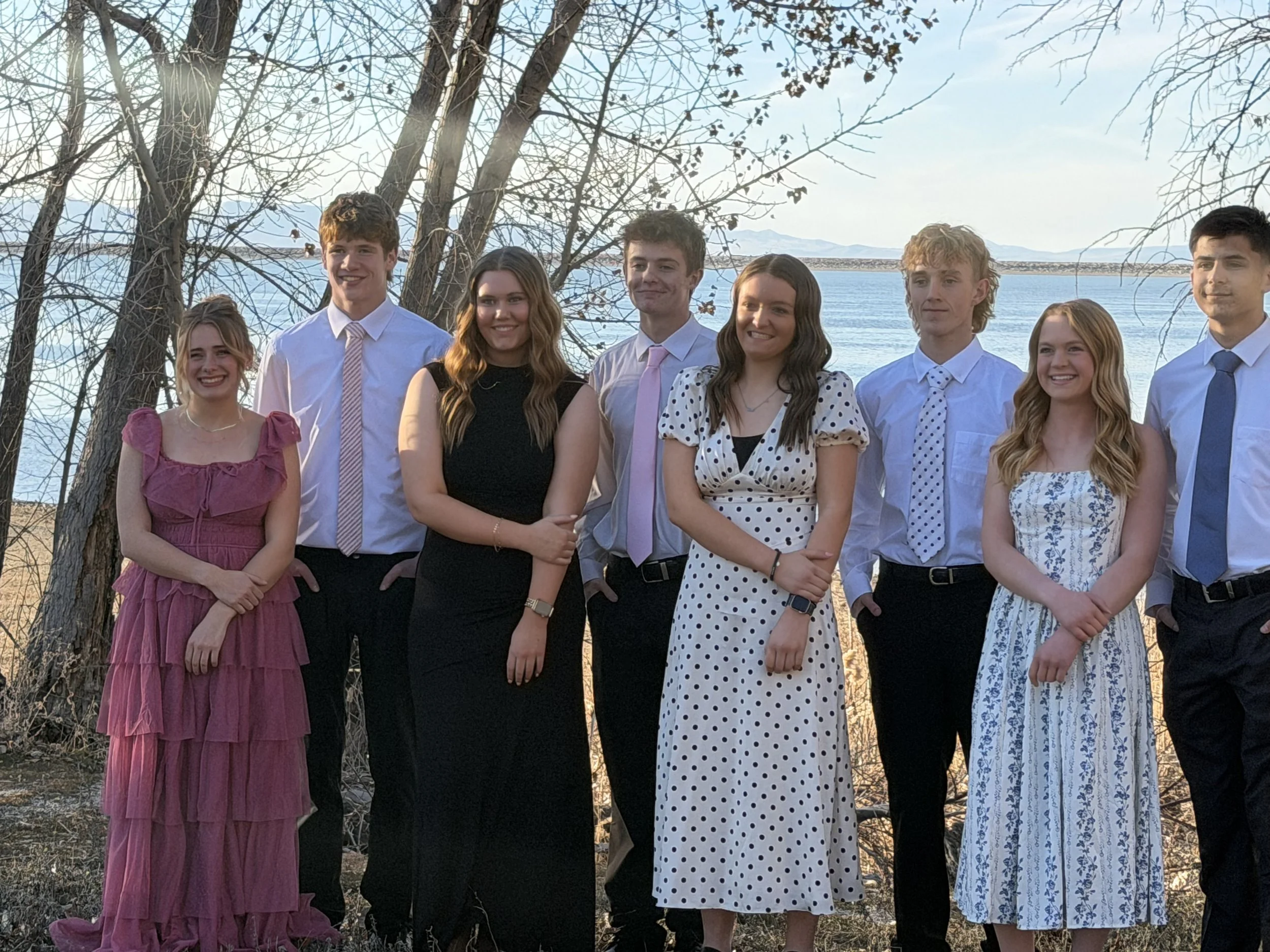 Group of nine young people outdoors near a body of water, smiling and dressed in semi-formal attire, with trees and a lake in the background.