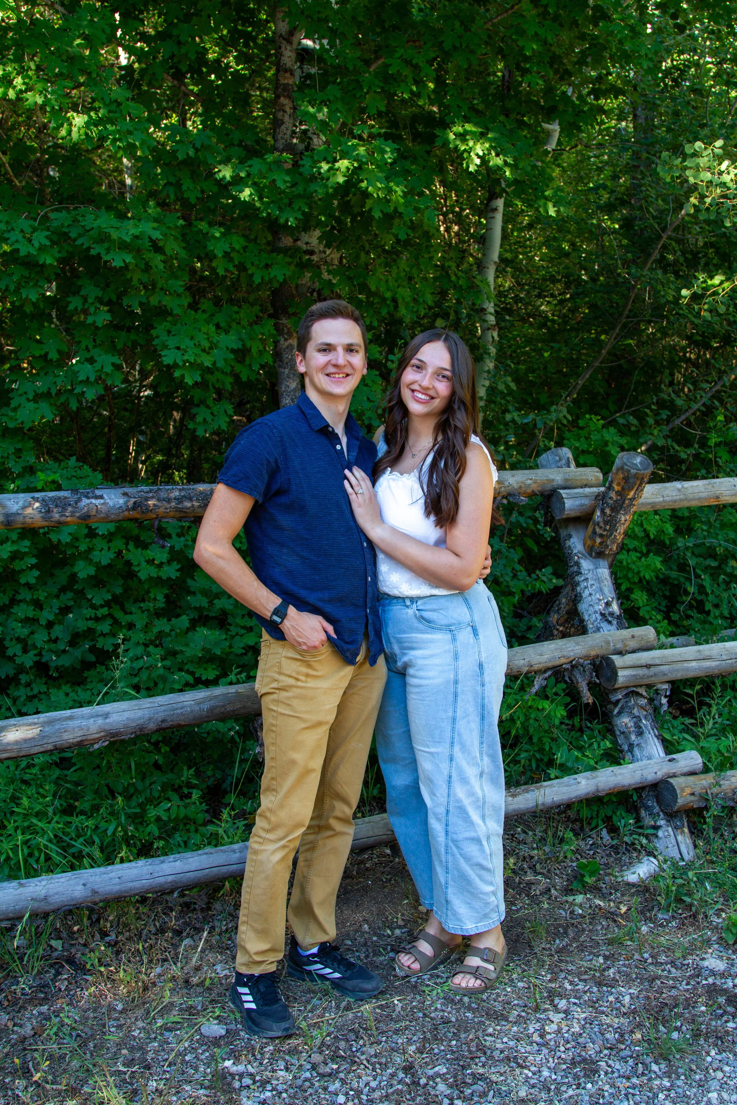 A young man and woman standing outdoors in front of a wooden fence and green foliage, smiling and embracing.