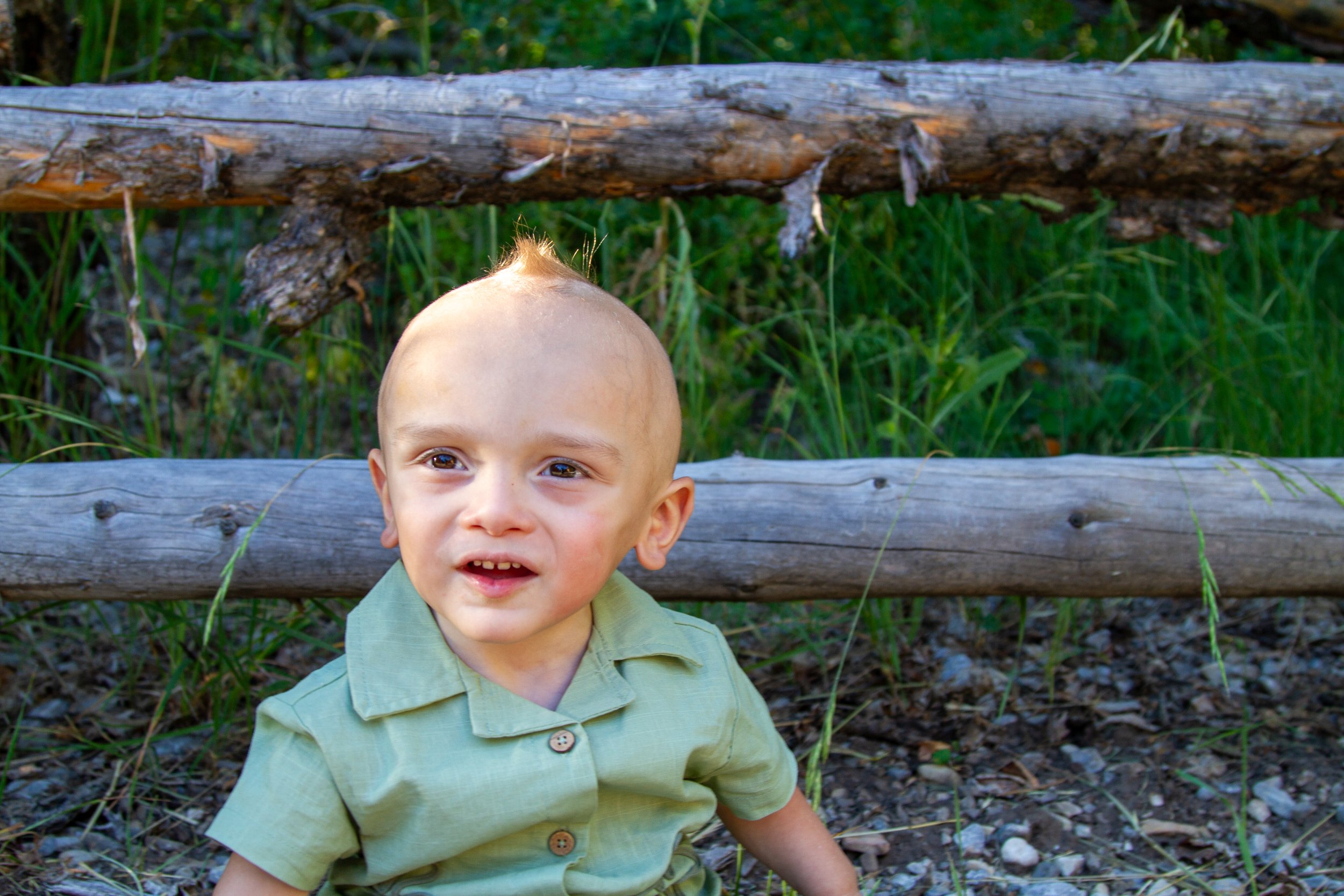 A young boy with a shaved head and green shirt smiling outdoors near a wooden fence with green plants in the background.