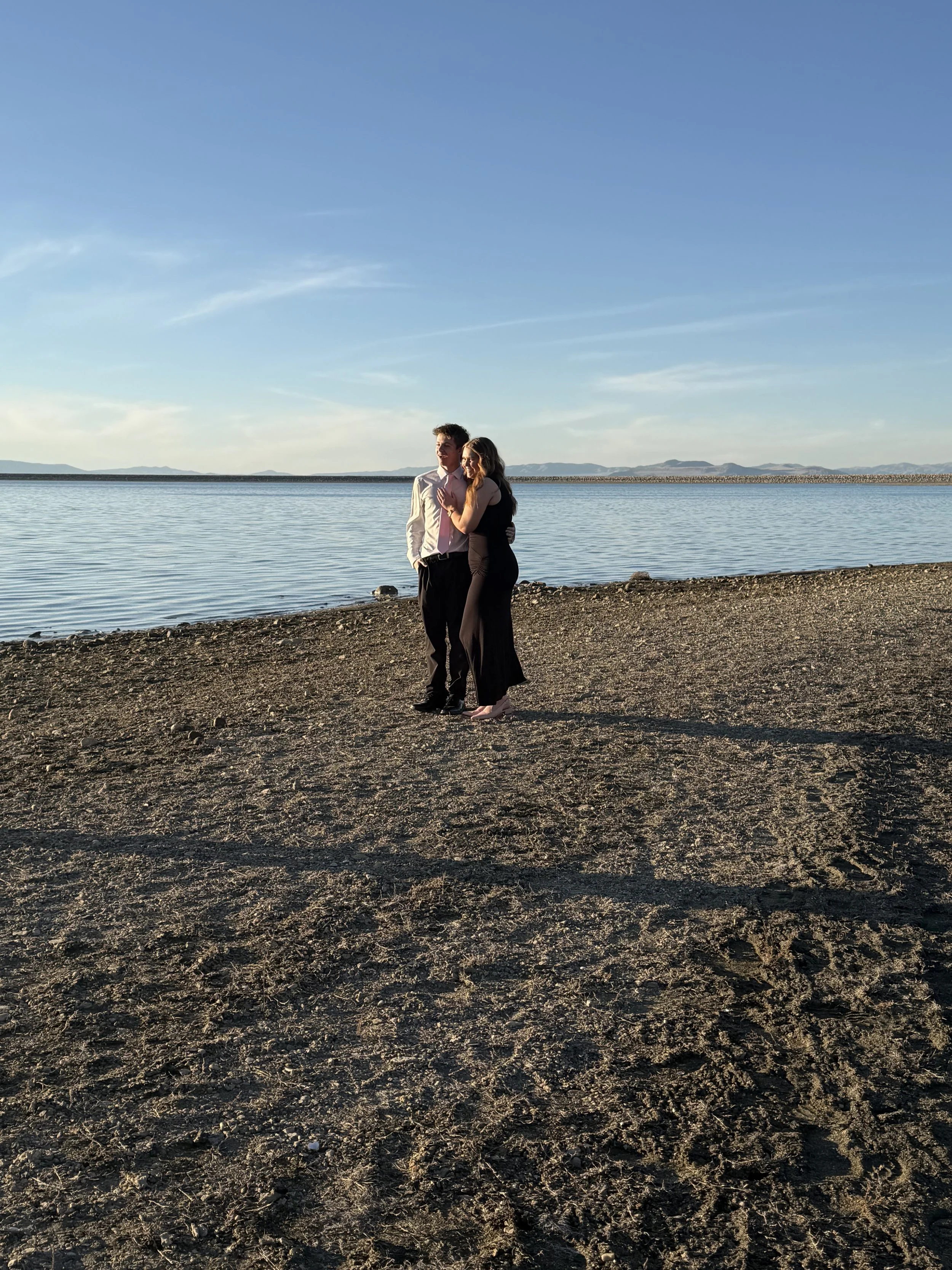 A couple stands on a sandy beach near a calm body of water, with mountains in the distance and a clear, blue sky overhead.