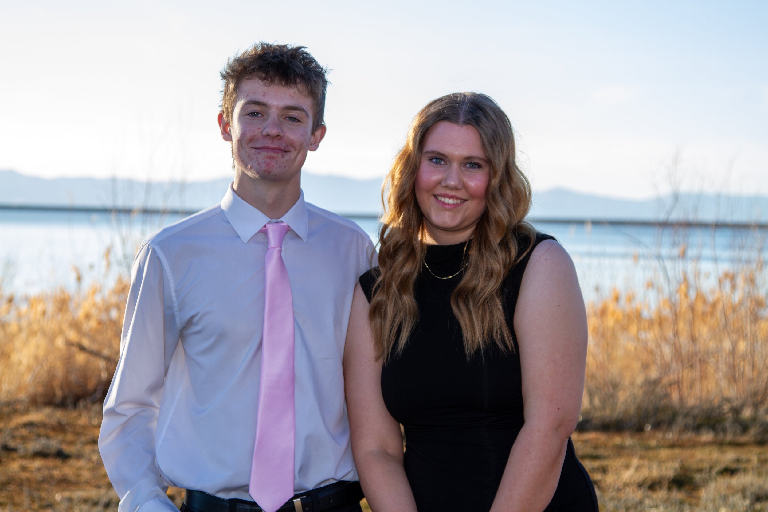 A young man and woman standing outdoors near a body of water with mountains in the background, smiling at the camera, during sunset.