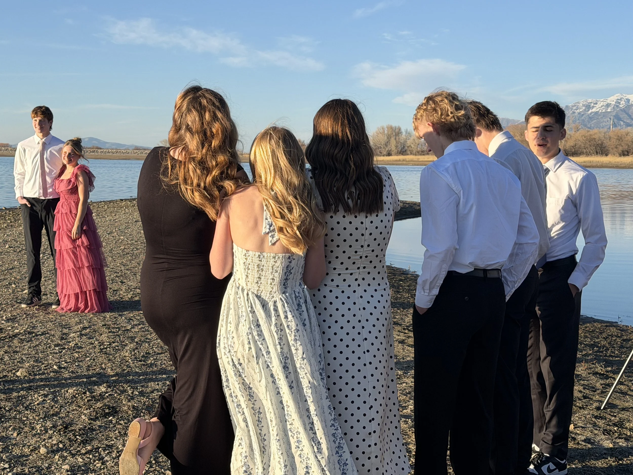 A group of teenagers dressed in formal attire are standing by a lake with mountains in the background, during sunset.