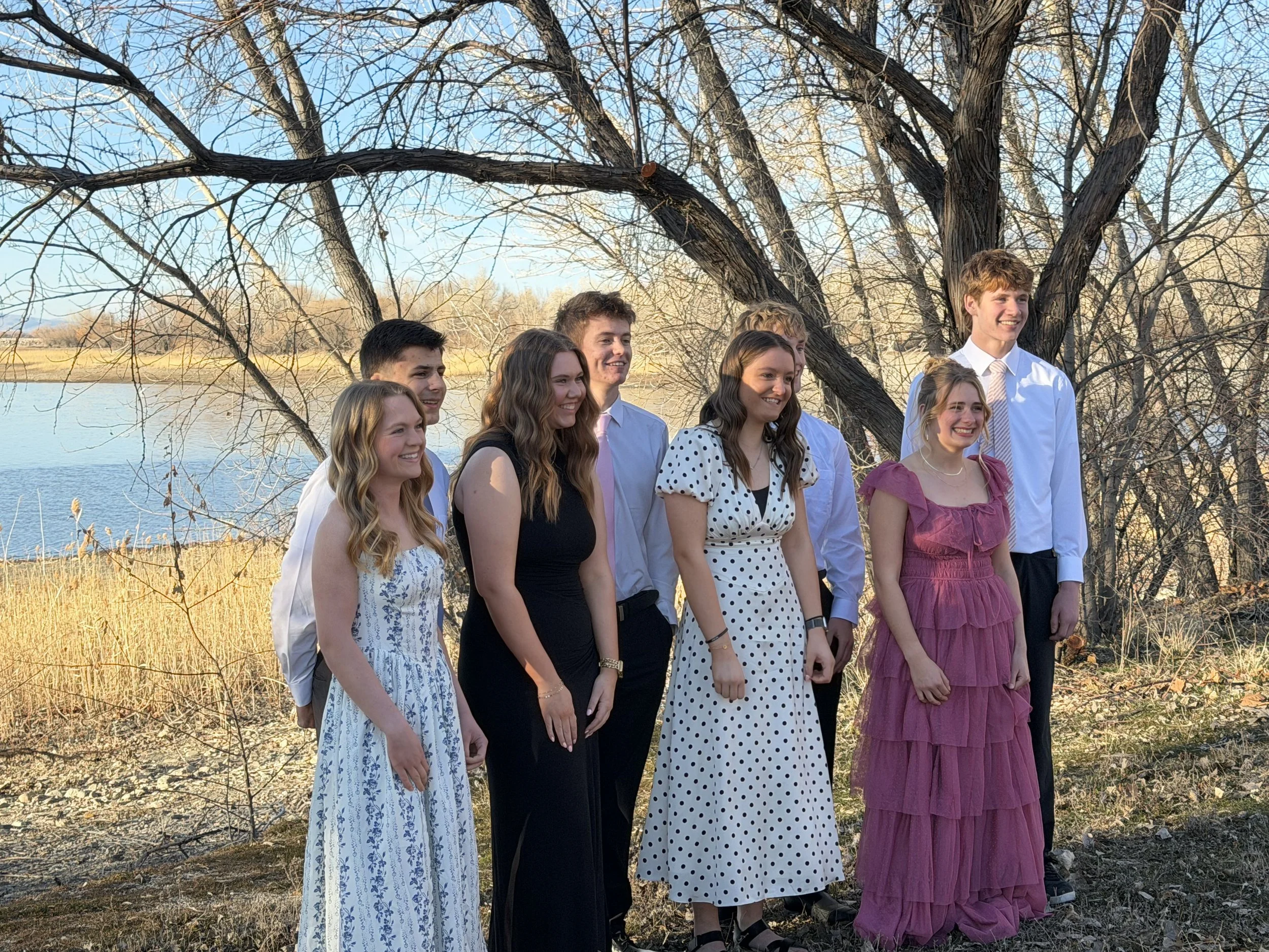 A group of nine young people, dressed in semi-formal attire, posing for a photo outdoors near a lake with trees in the background.