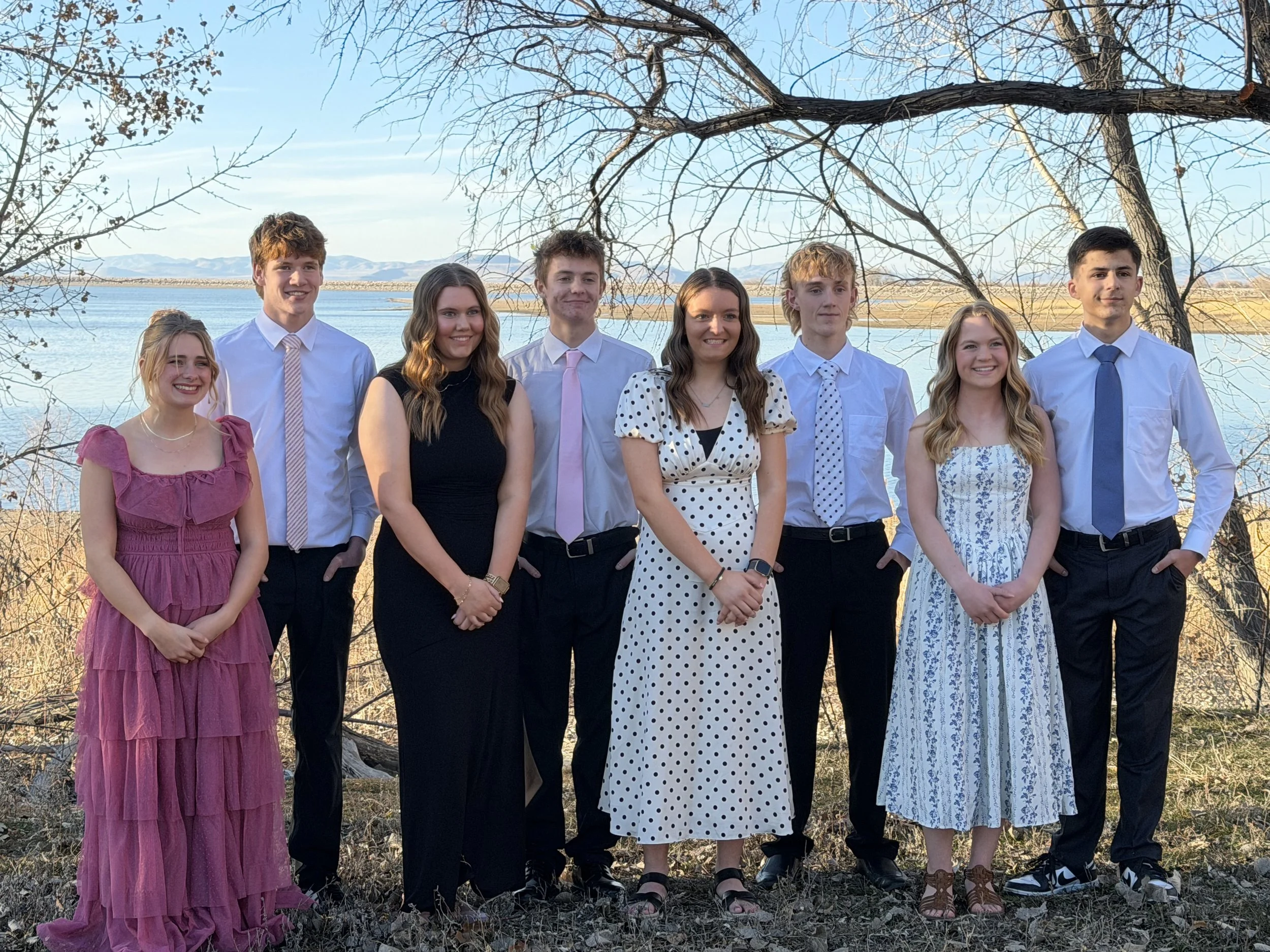 A group of eight young people posing outdoors near a lake with trees and mountains in the background, dressed in semi-formal attire.