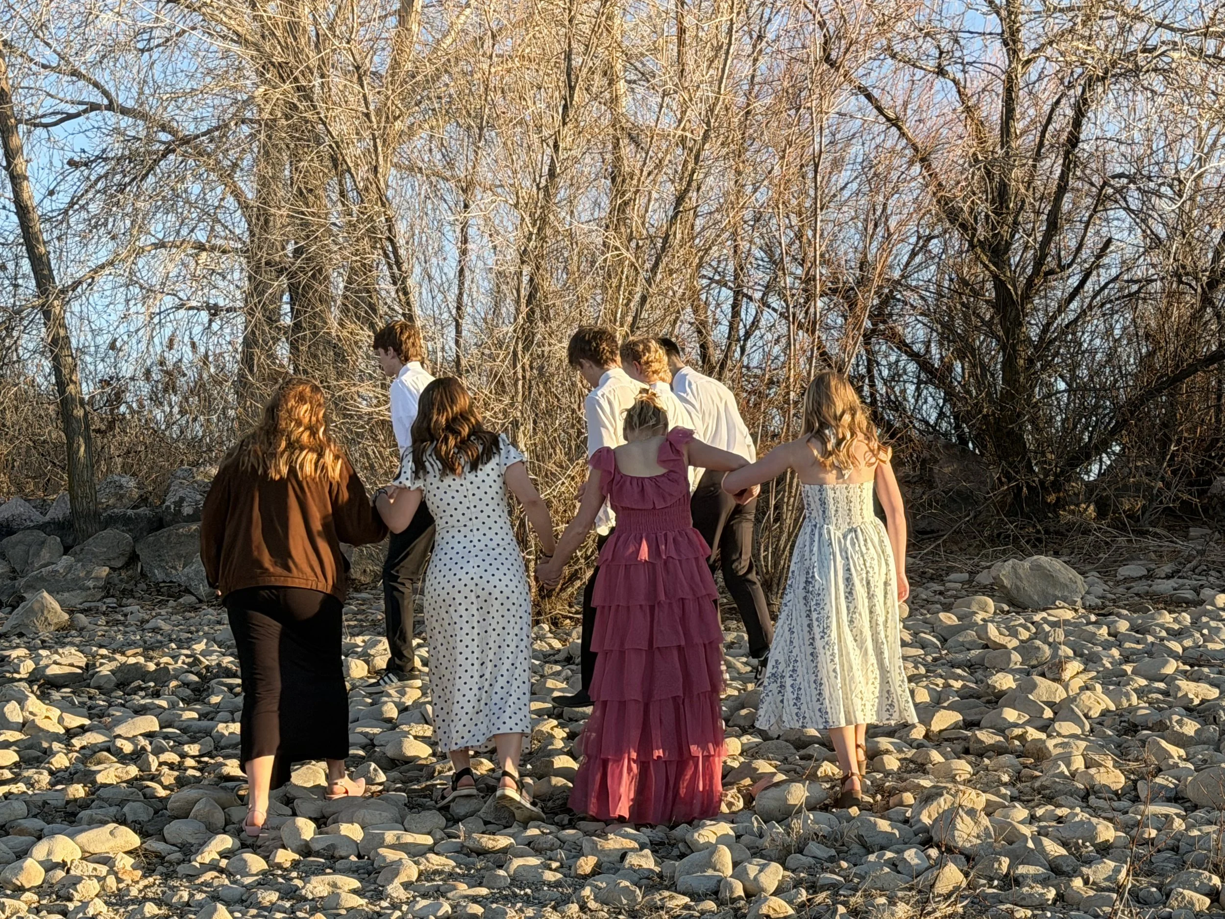Group of women and men holding hands and walking in a circle outdoors on rocky ground, with leafless trees in the background and warm sunlight.
