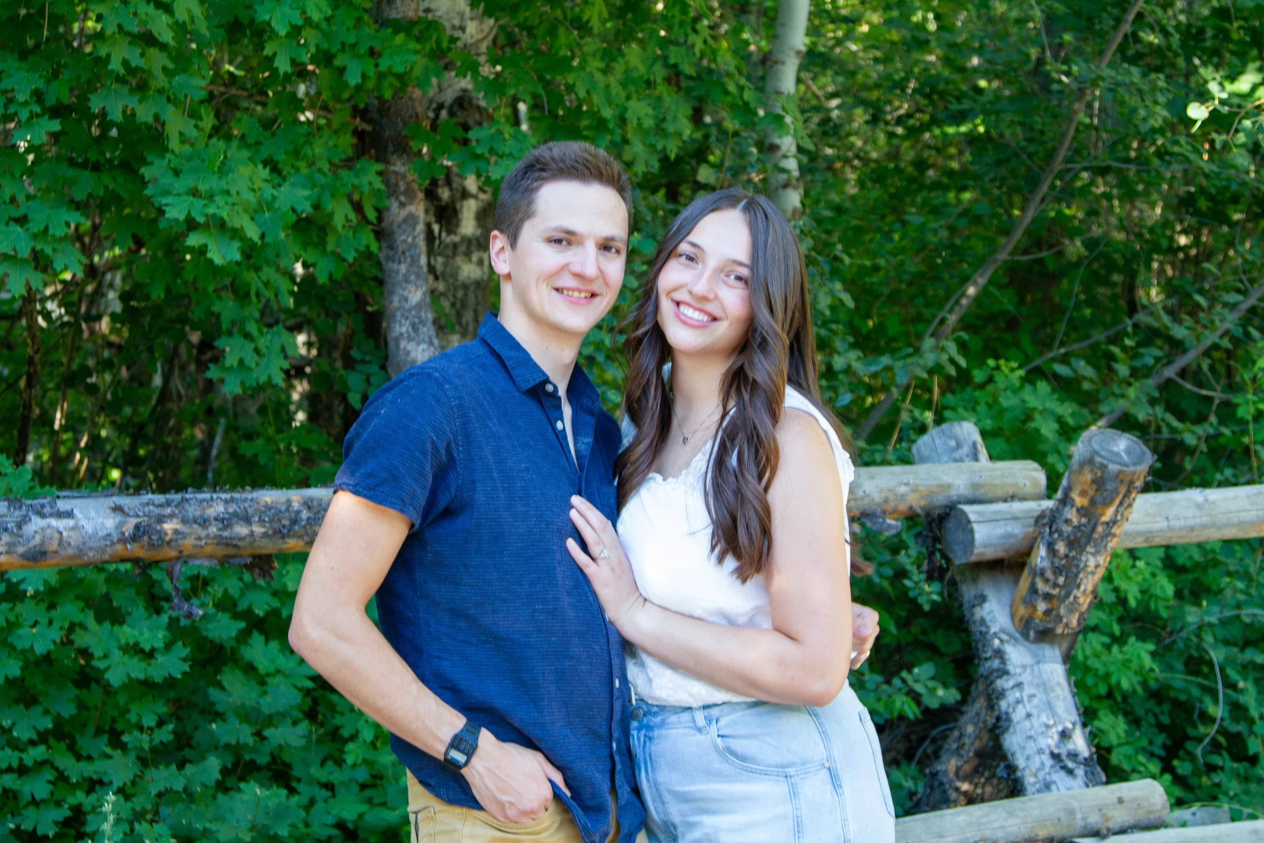 A smiling young man and woman standing close together outdoors, with lush green trees and a wooden fence in the background.