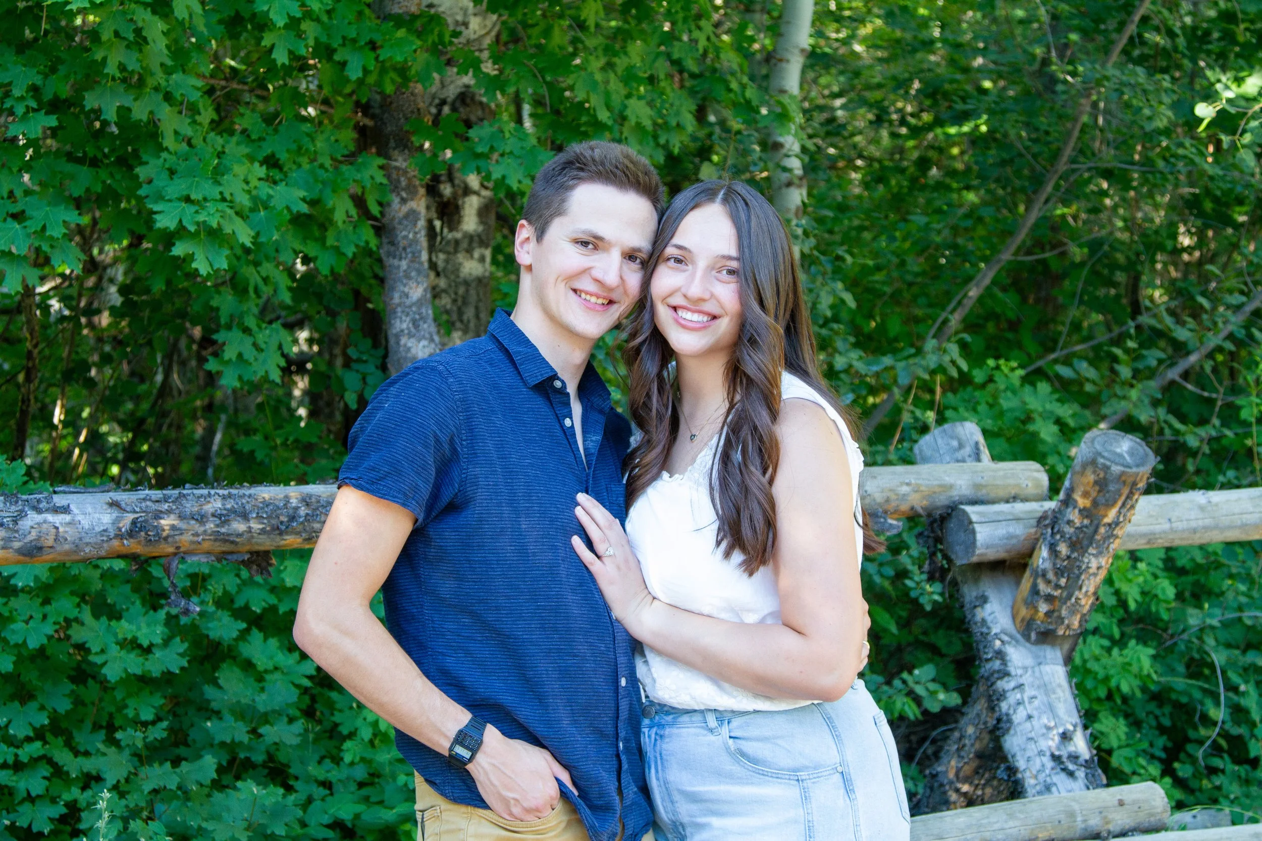 A young man and woman are posing together outdoors, smiling and standing close to each other, with a background of green trees and a rustic wooden fence.
