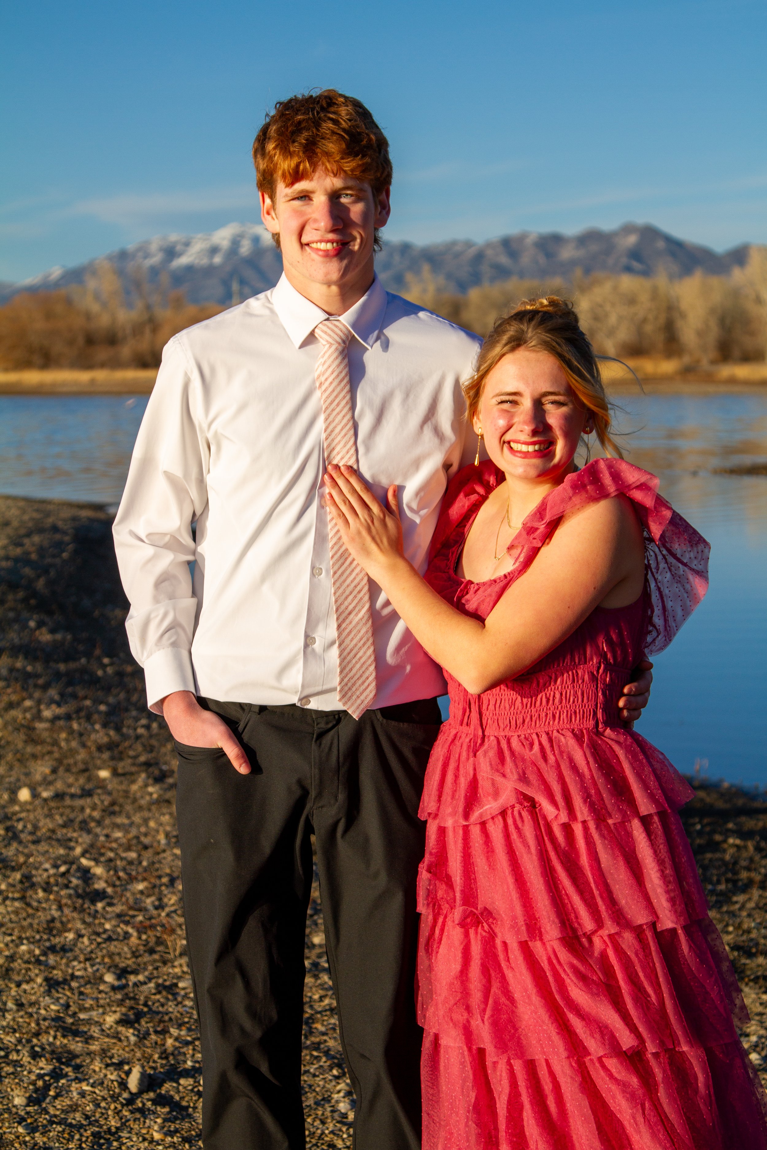 A young man in a white shirt and tie and a young woman in a pink dress smiling and posing together by a river with mountains in the background during sunset.