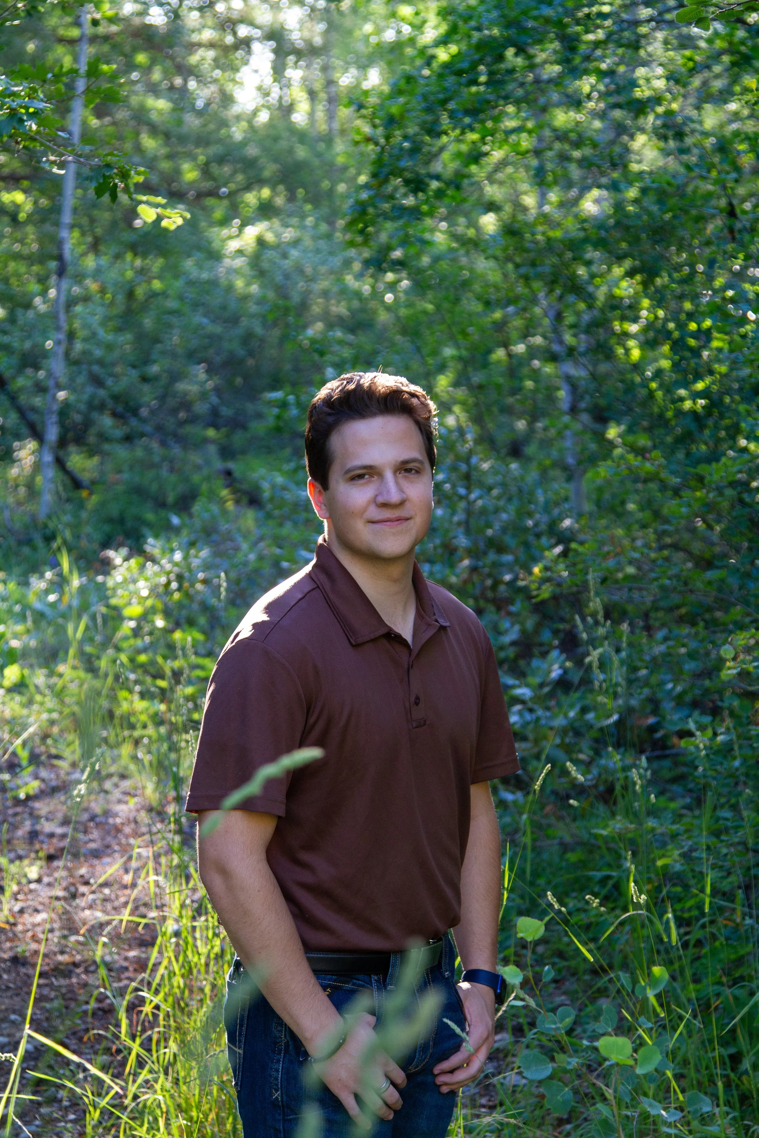 A young man standing outdoors in a wooded area with green foliage and sunlight filtering through trees.