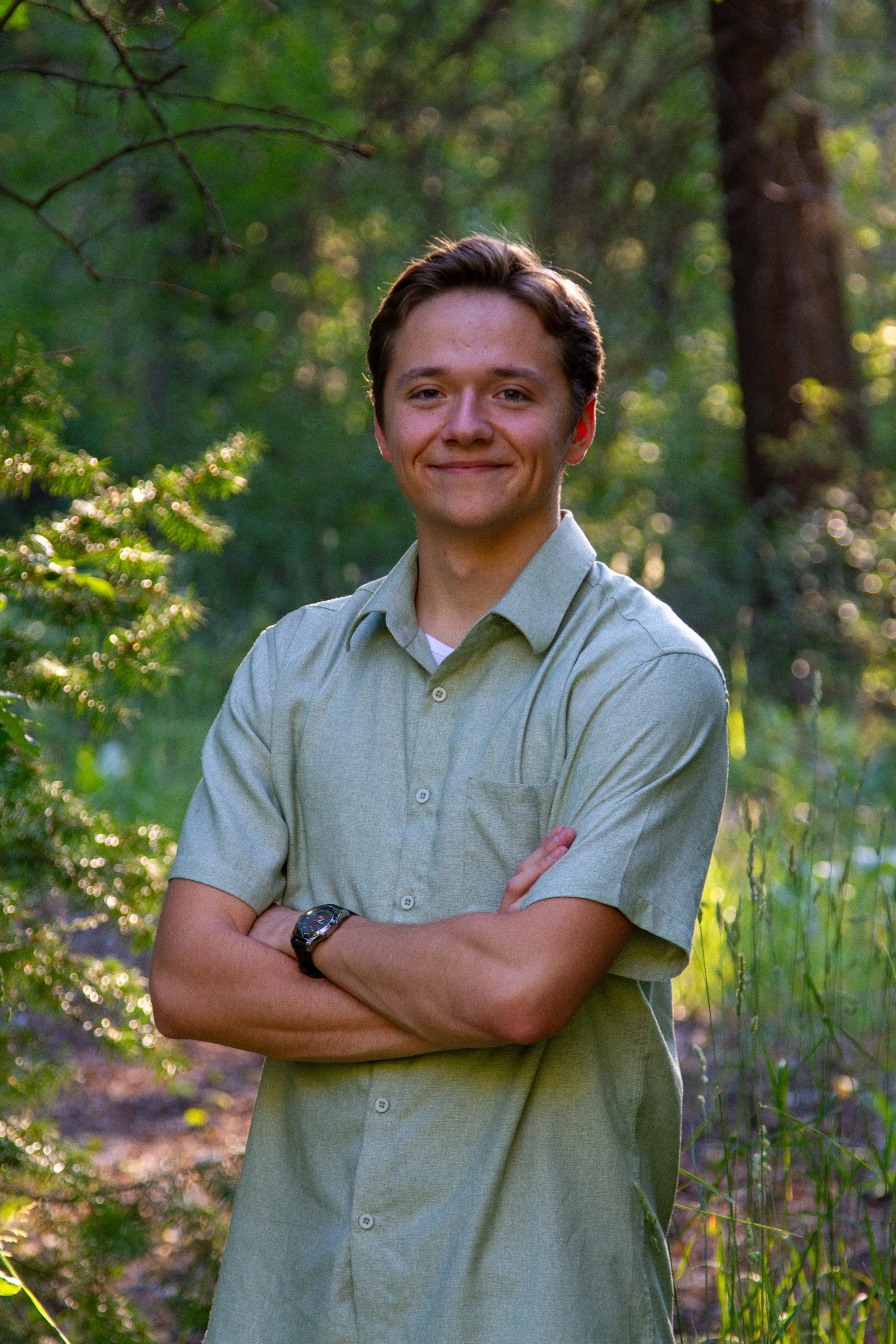 A young man with crossed arms, smiling, wearing a light green button-up shirt, standing outdoors in a forest with sunlight filtering through the trees.