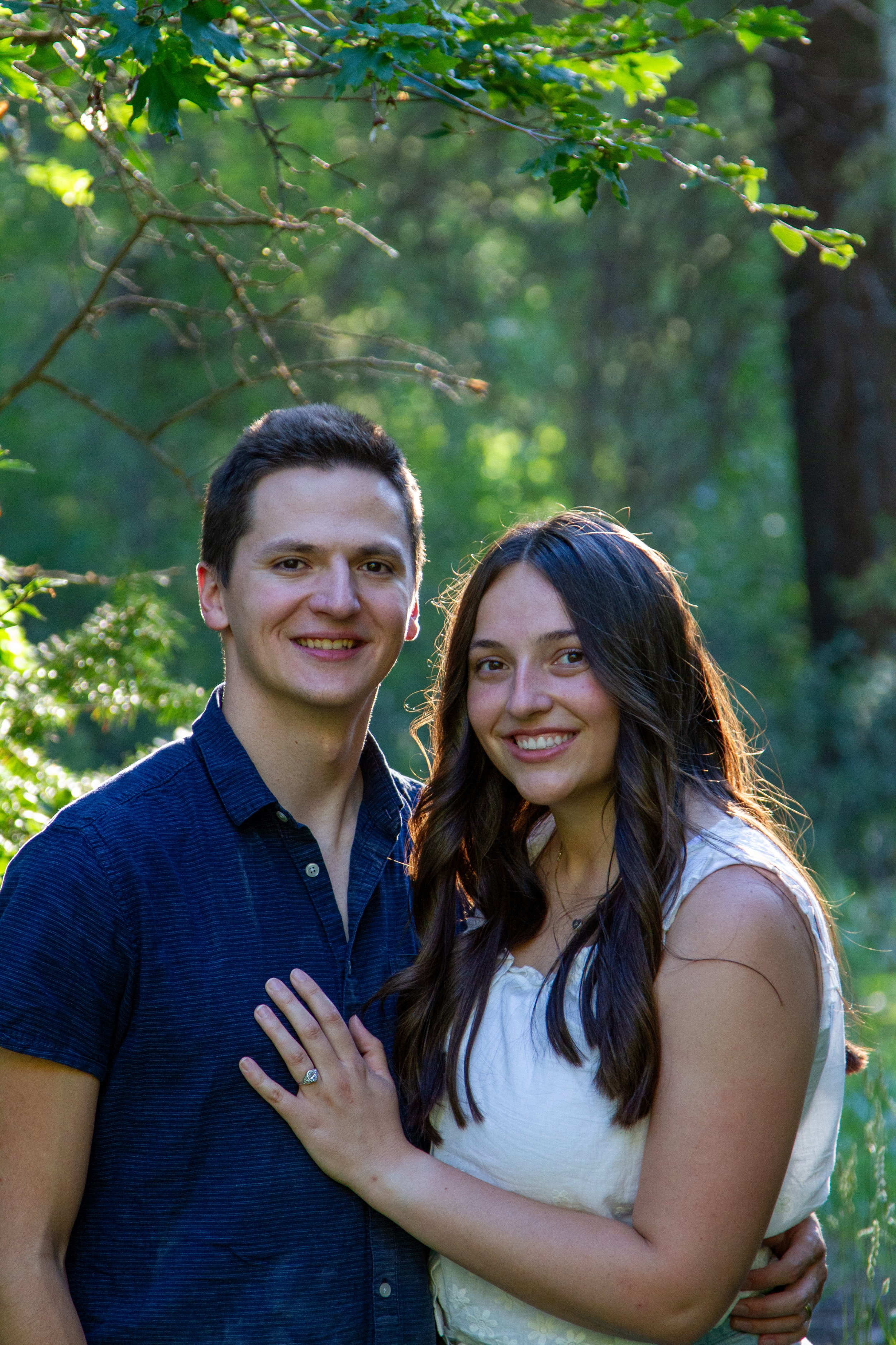 A smiling man and woman standing close together outdoors with trees and greenery in the background. The woman is wearing a white sleeveless top, and the man is wearing a dark blue shirt. The woman's left arm is resting on the man's chest, and she has