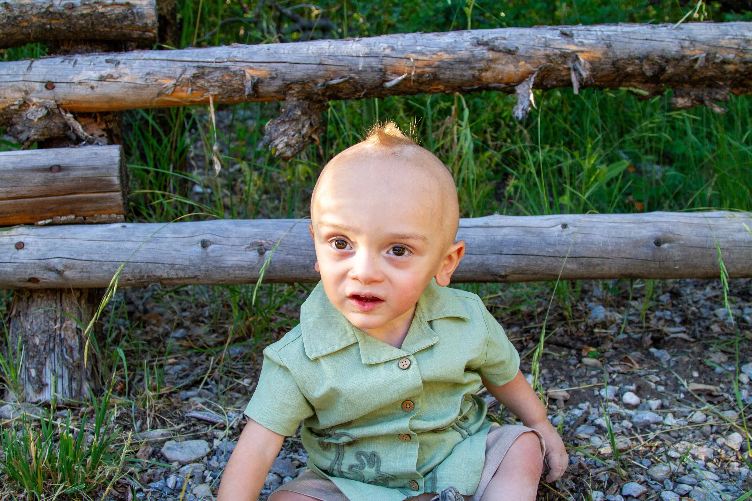 A young boy with a bald head sitting outdoors on a rocky ground near a wooden fence, with greenery in the background.