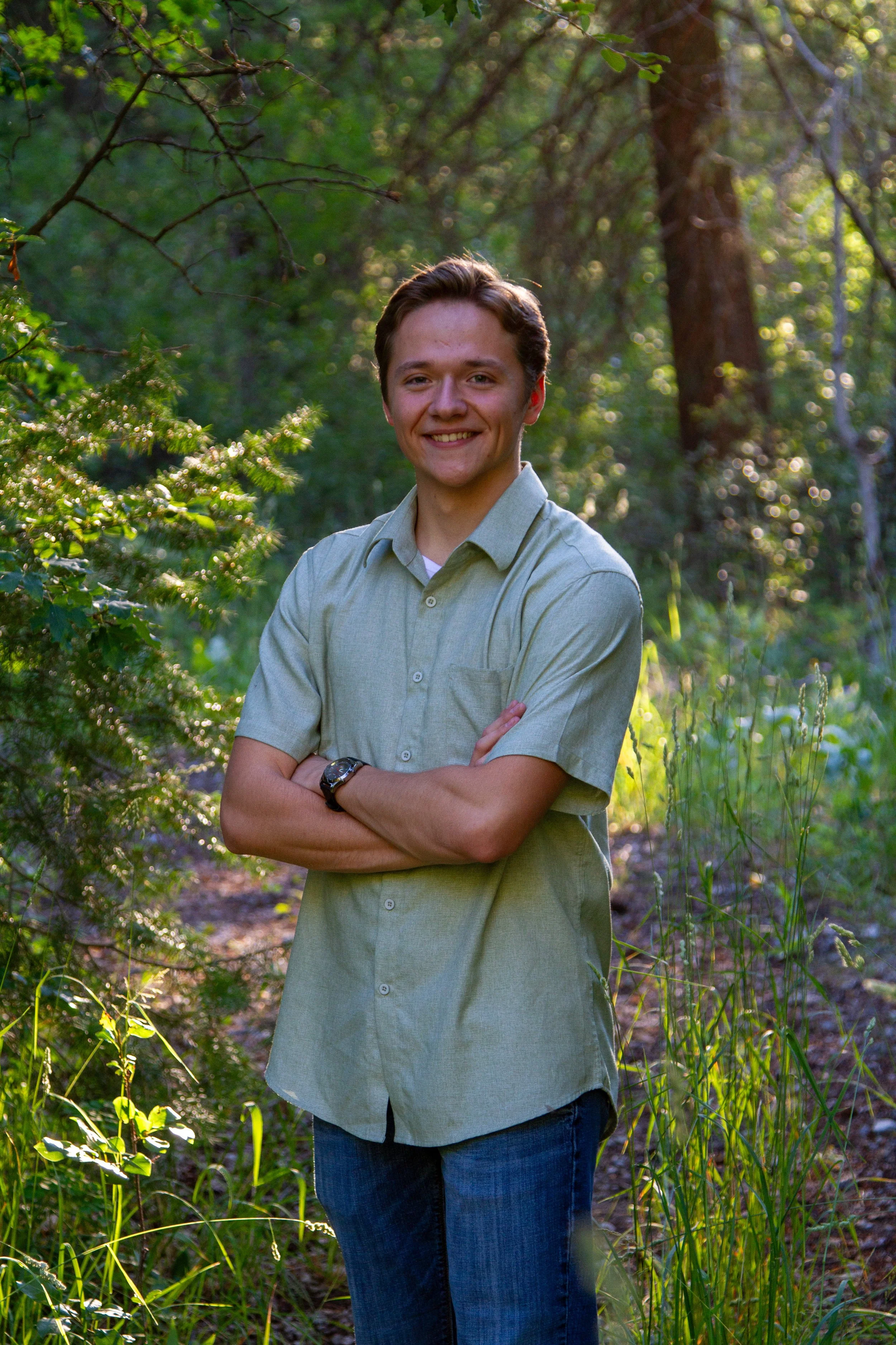 A young man with light brown hair, wearing a light green button-up shirt and dark blue jeans, standing outdoors in a forest with lush green foliage and sunlight filtering through trees, smiling at the camera with arms crossed.