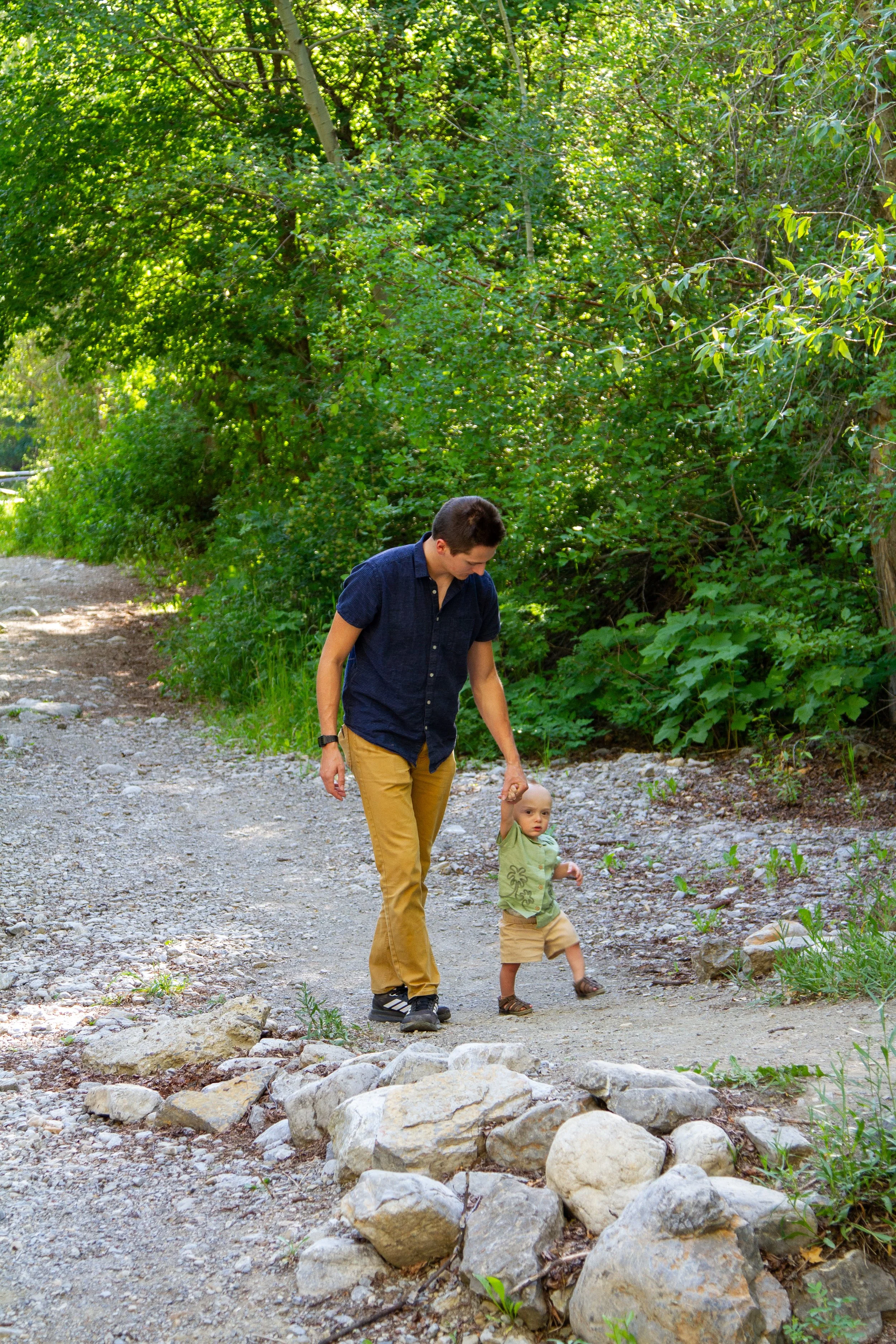 A man and a young boy walking on a rocky dirt path in a green forested area. The man is holding the boy's hand as they walk.