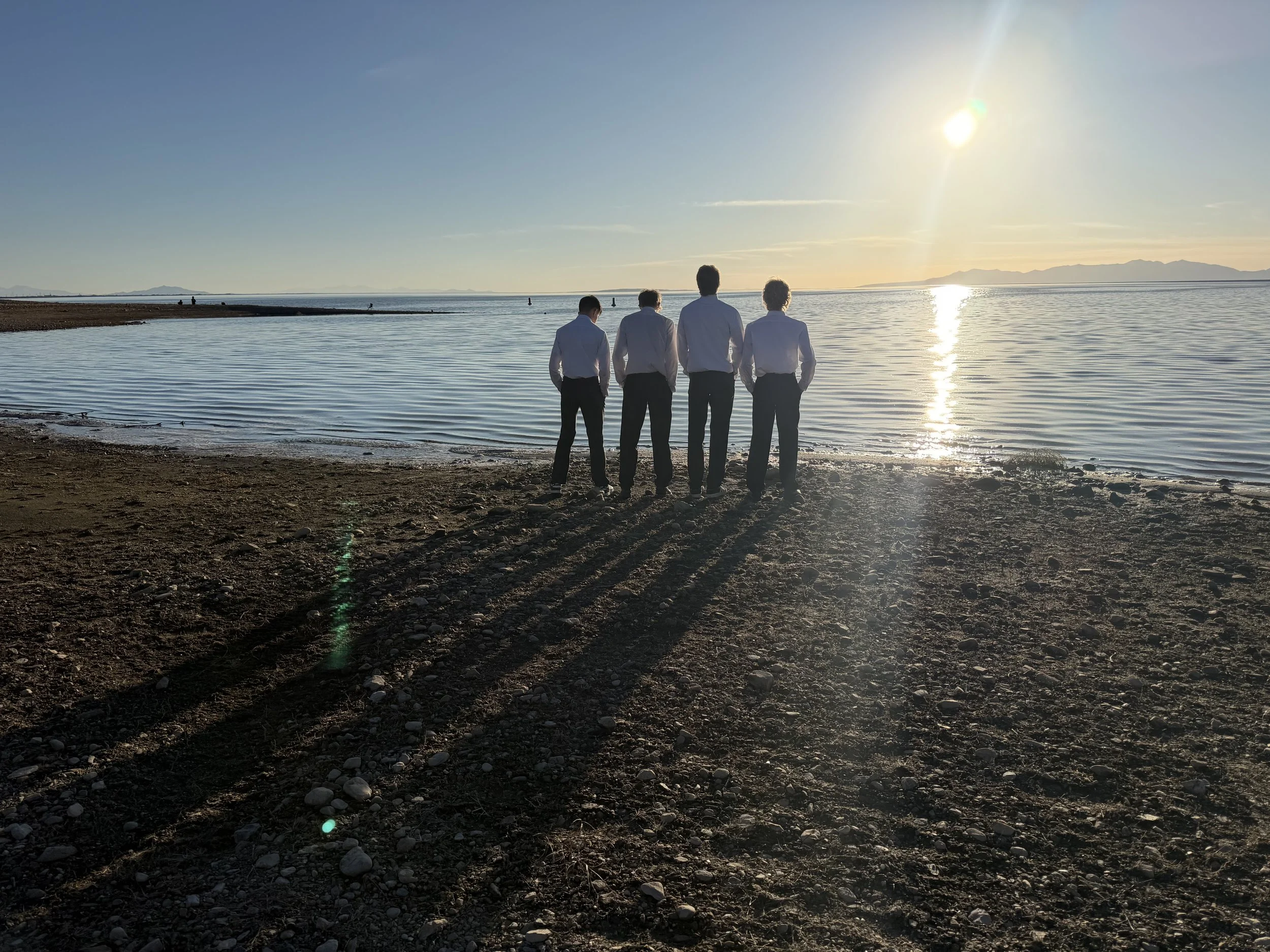Four men in dress shirts and pants standing on a beach at sunset, facing the water and casting long shadows.
