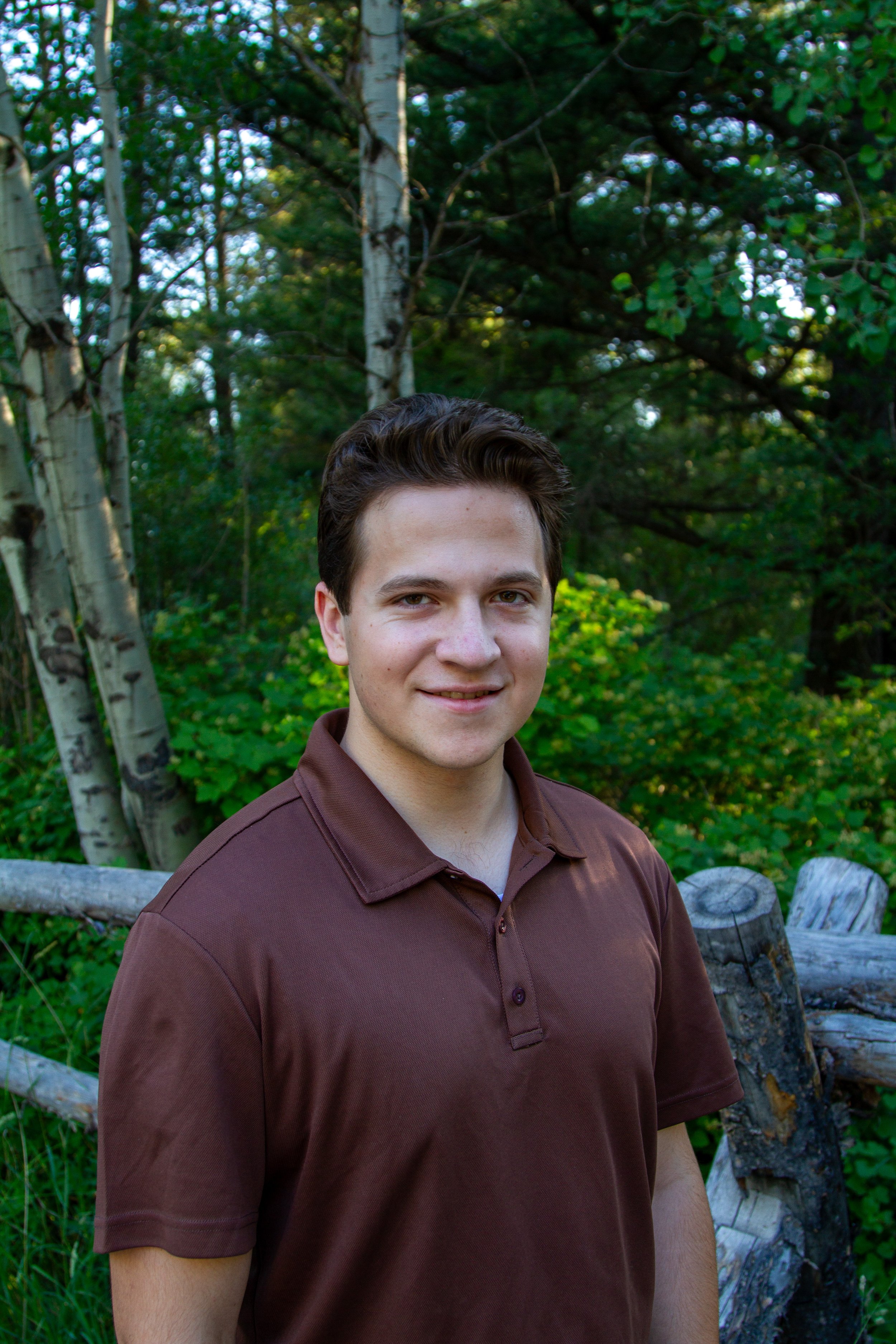 A young man with short dark hair, wearing a brown polo shirt, standing outdoors with trees and greenery in the background.