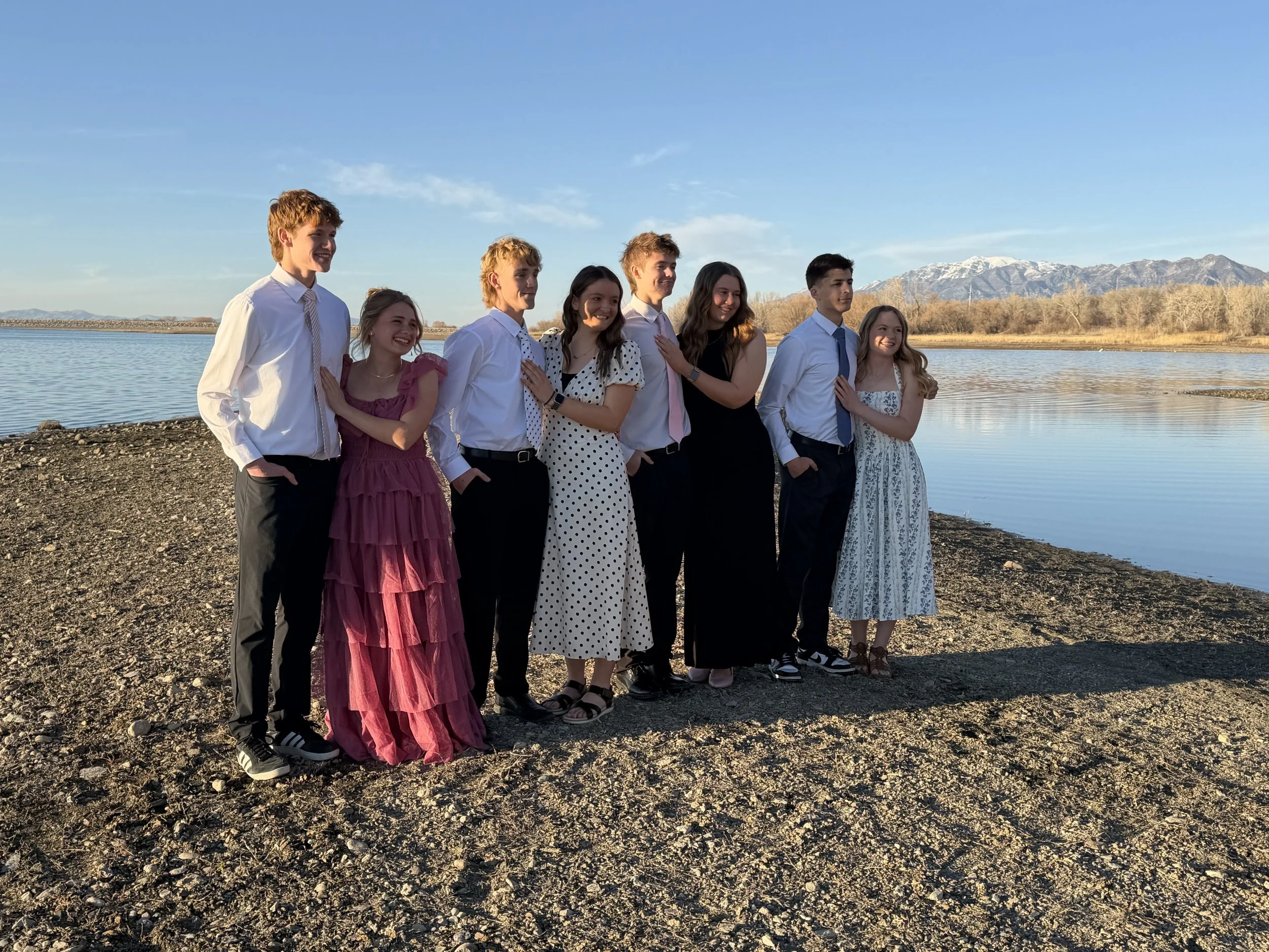 Group of nine teenagers standing near a body of water with mountains in the background, dressed in semi-formal attire.