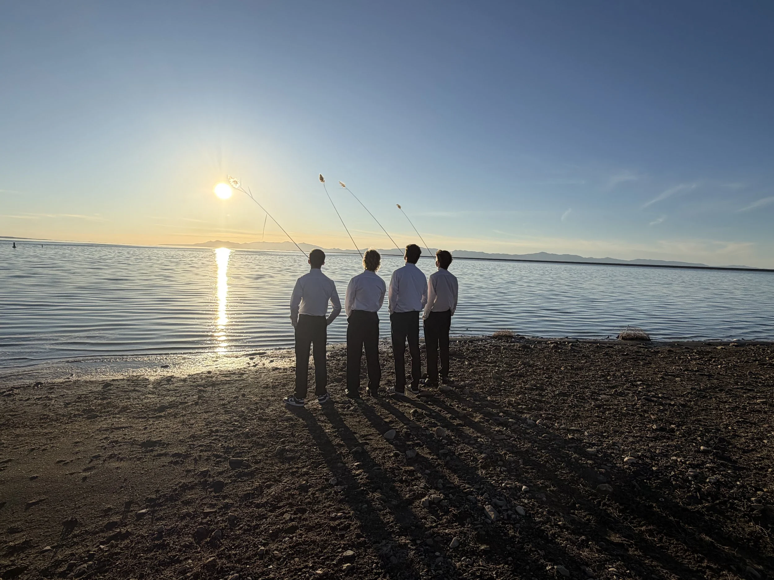 Four men in business attire standing on a rocky beach at sunset, fishing with their lines cast into a calm body of water with mountains in the distance.