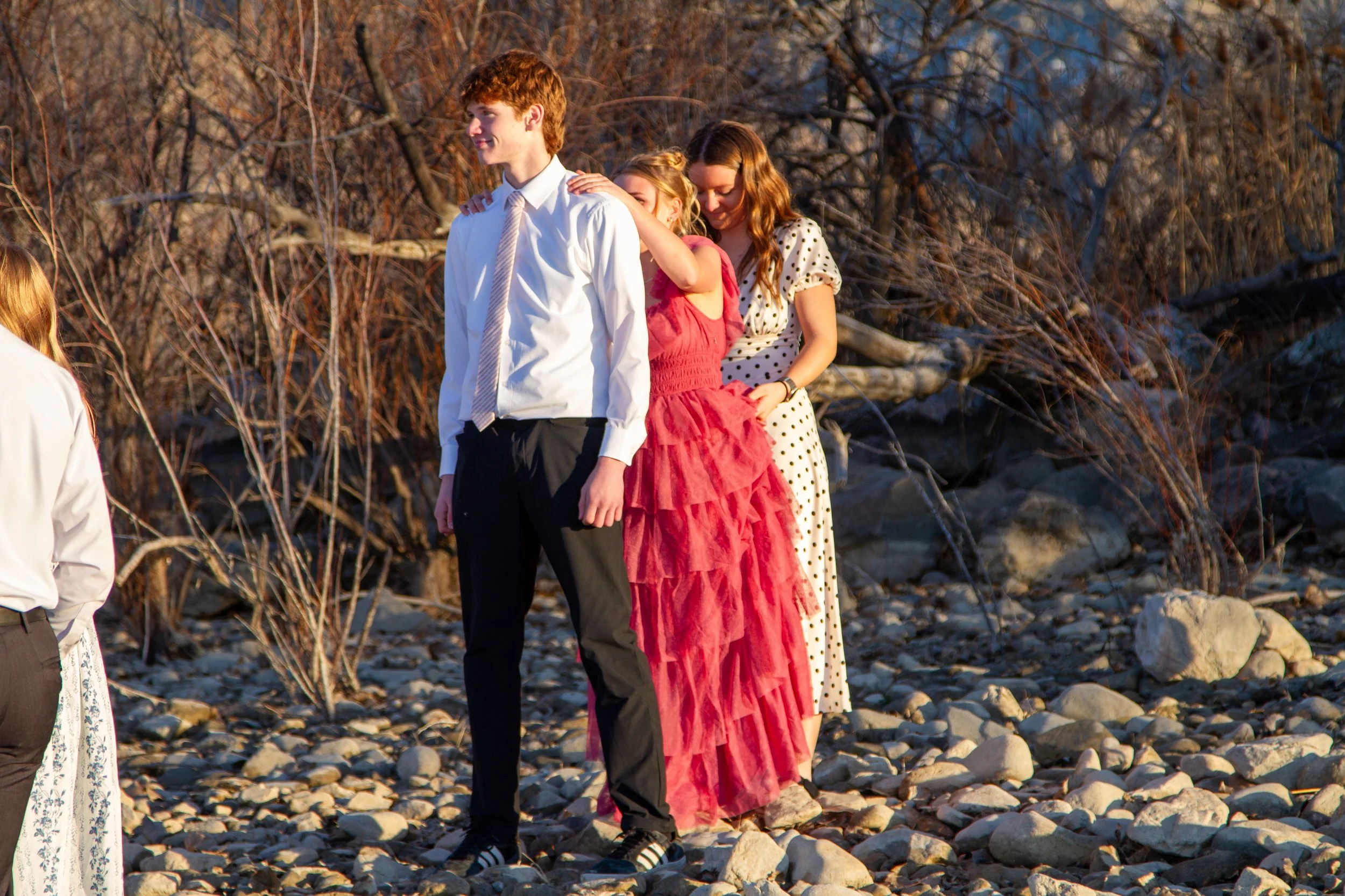 A group of four children on a rocky beach during sunset, with a background of leafless trees, one girl in a red dress is leaning on a tall boy in a white shirt and black pants, another girl in a polka dot dress is standing behind them, and a fourth g