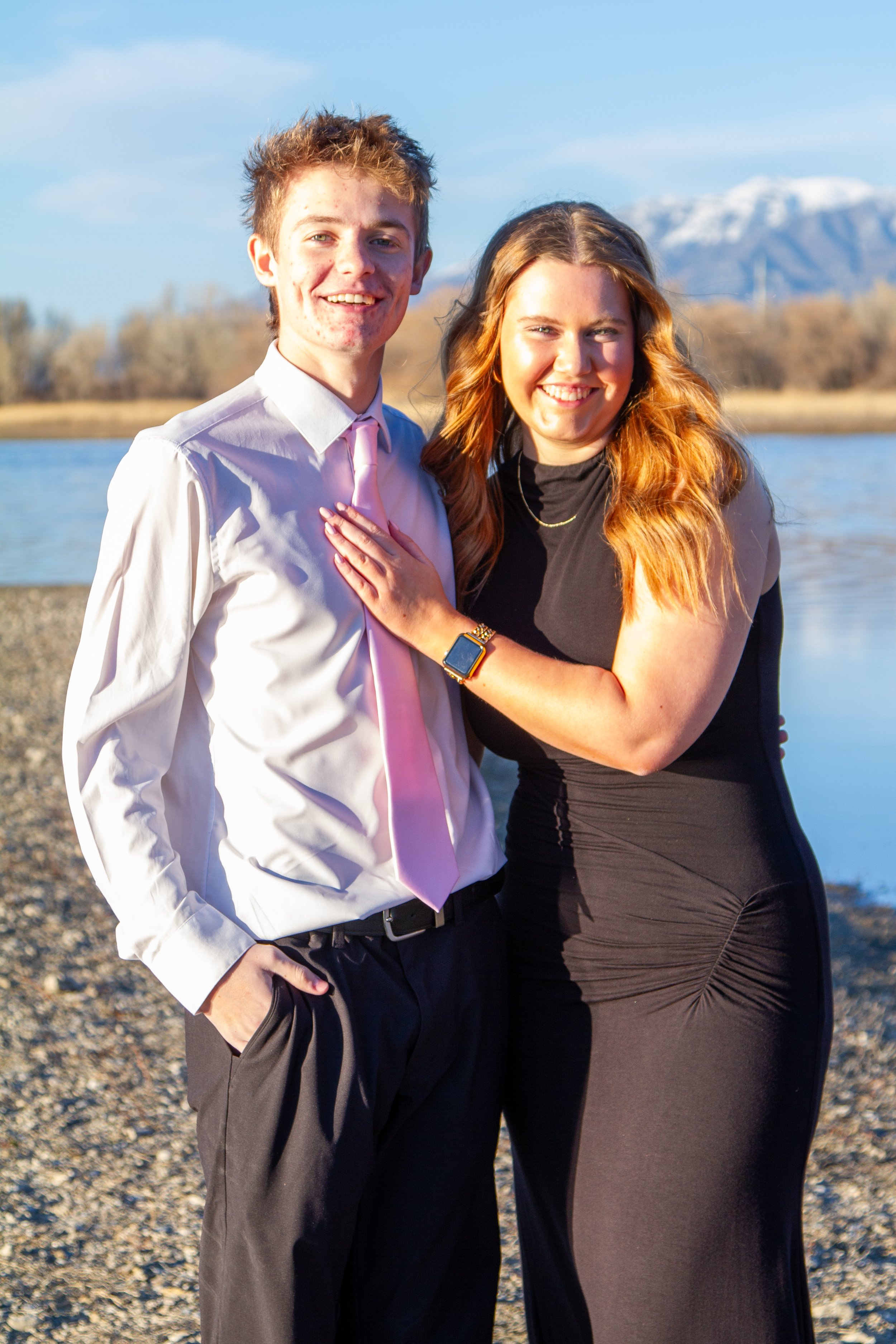 A smiling couple standing outdoors by a lake with snow-capped mountains in the background, during a sunny day.