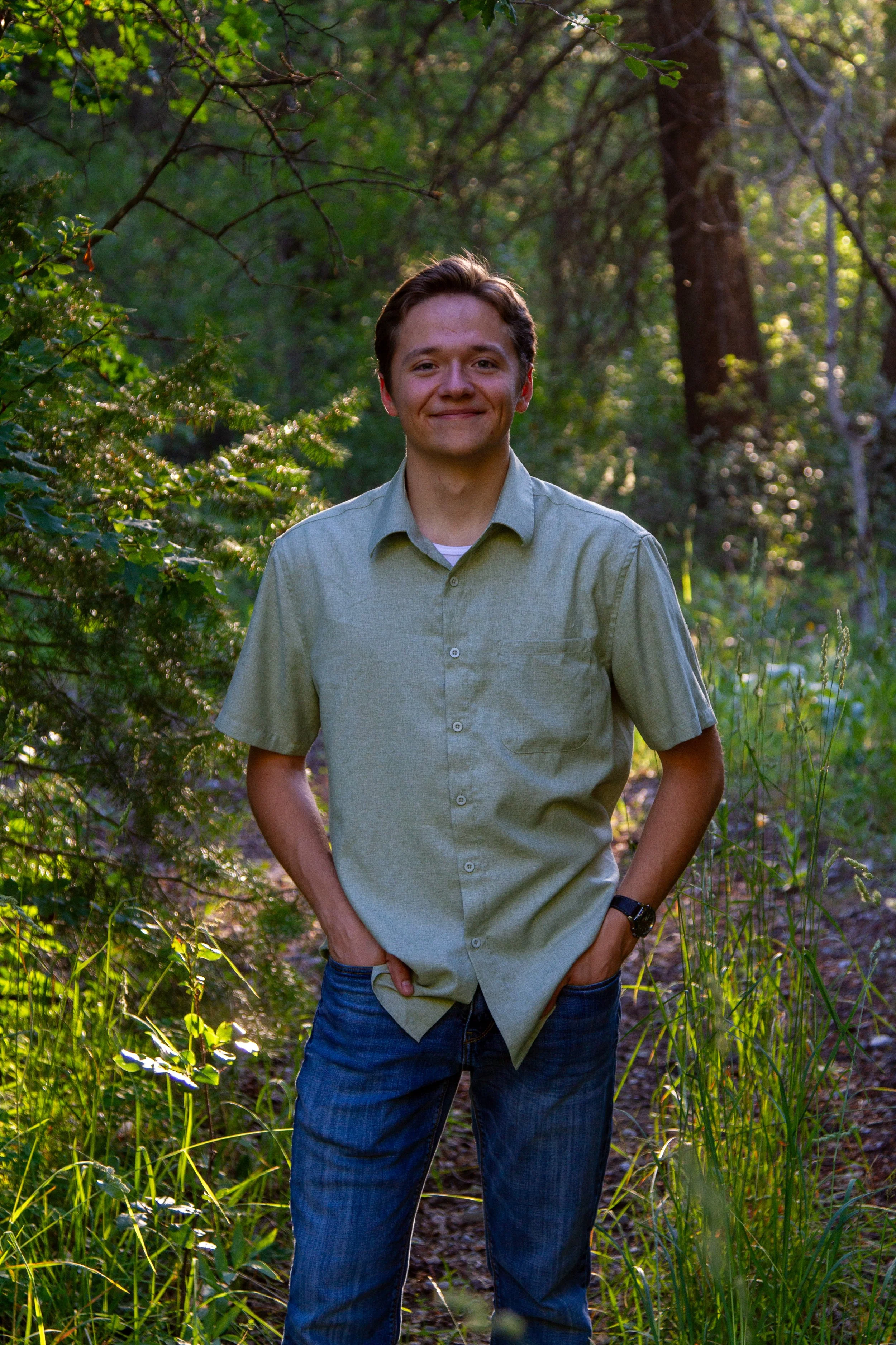 A young man with brown hair and a light smile, wearing a green button-up shirt and blue jeans, standing outdoors in a wooded area with sunlight filtering through the trees.