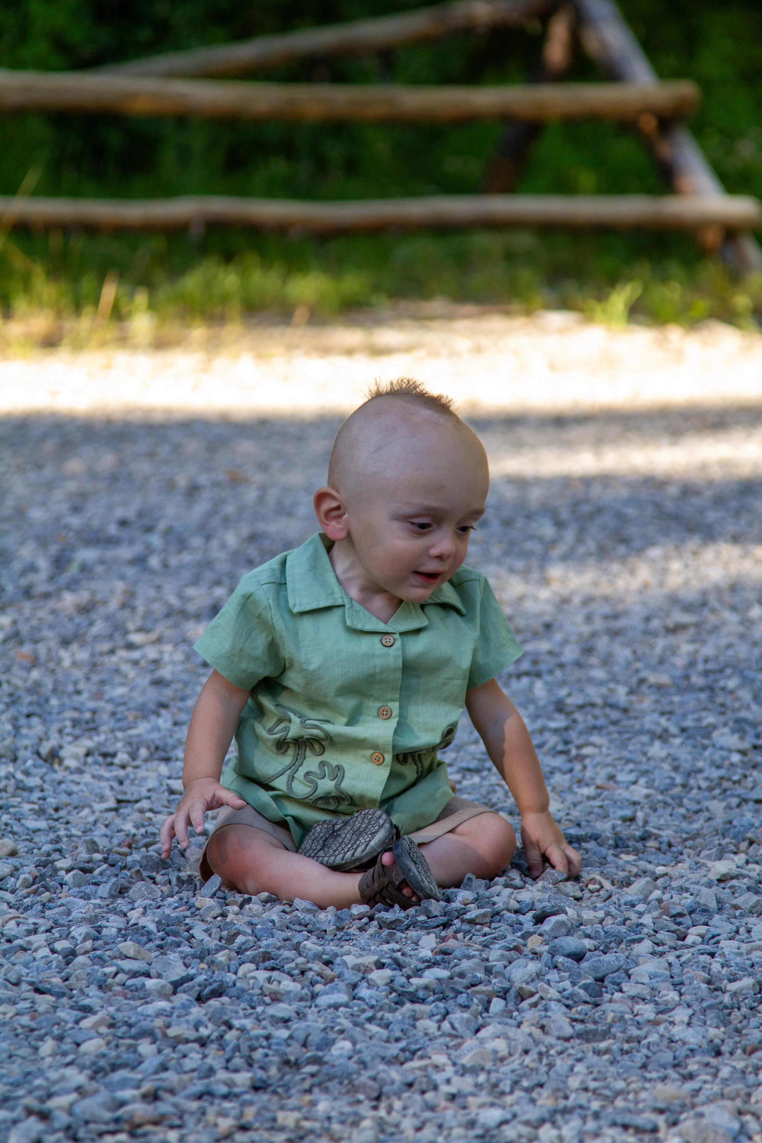 A young boy with light skin, a shaved head, wearing a green shirt and shorts, sitting on a rocky surface outdoors, with a blurred background of trees and a wooden structure.