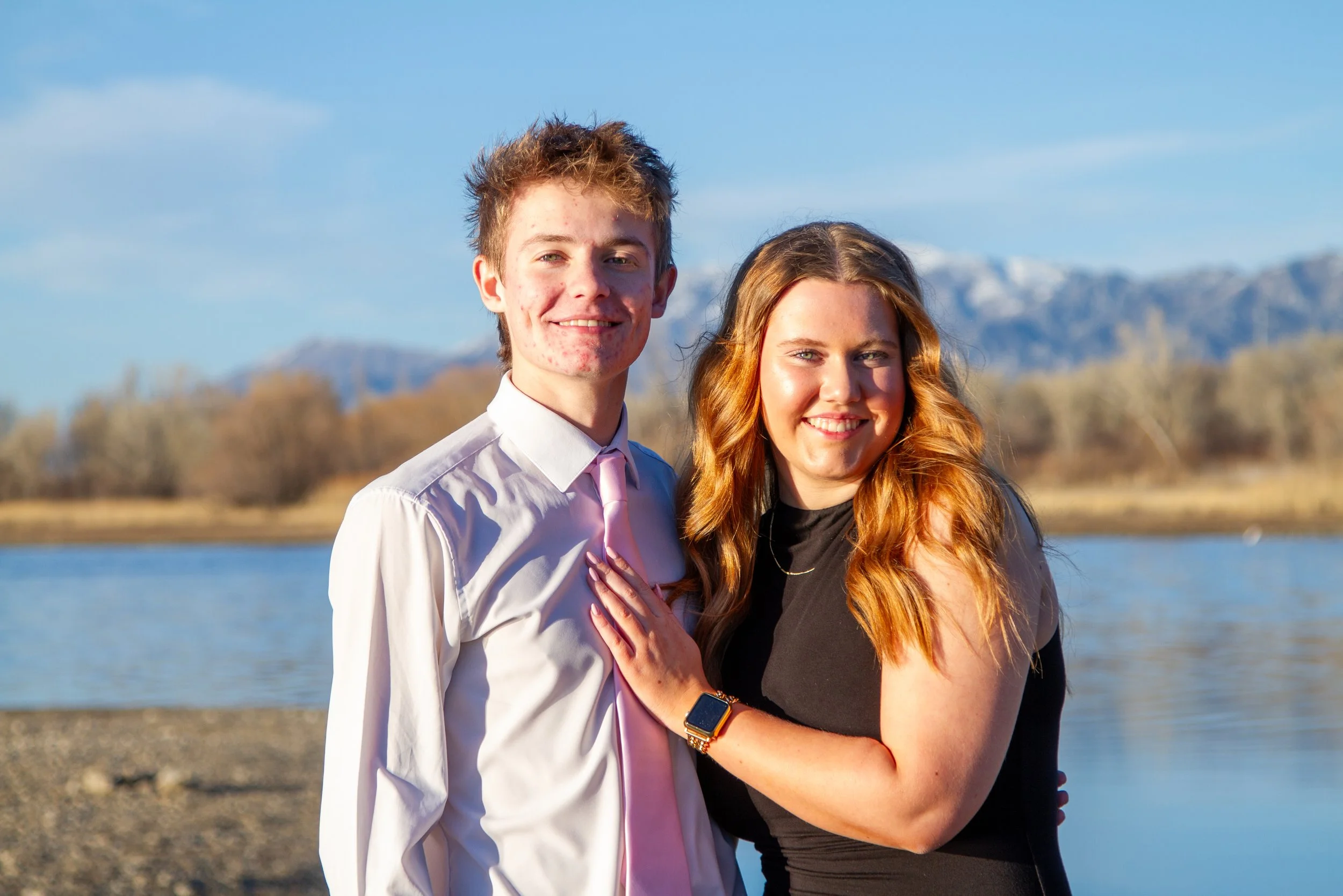 A young man and woman smiling outdoors near a body of water with mountains in the background, during daytime.